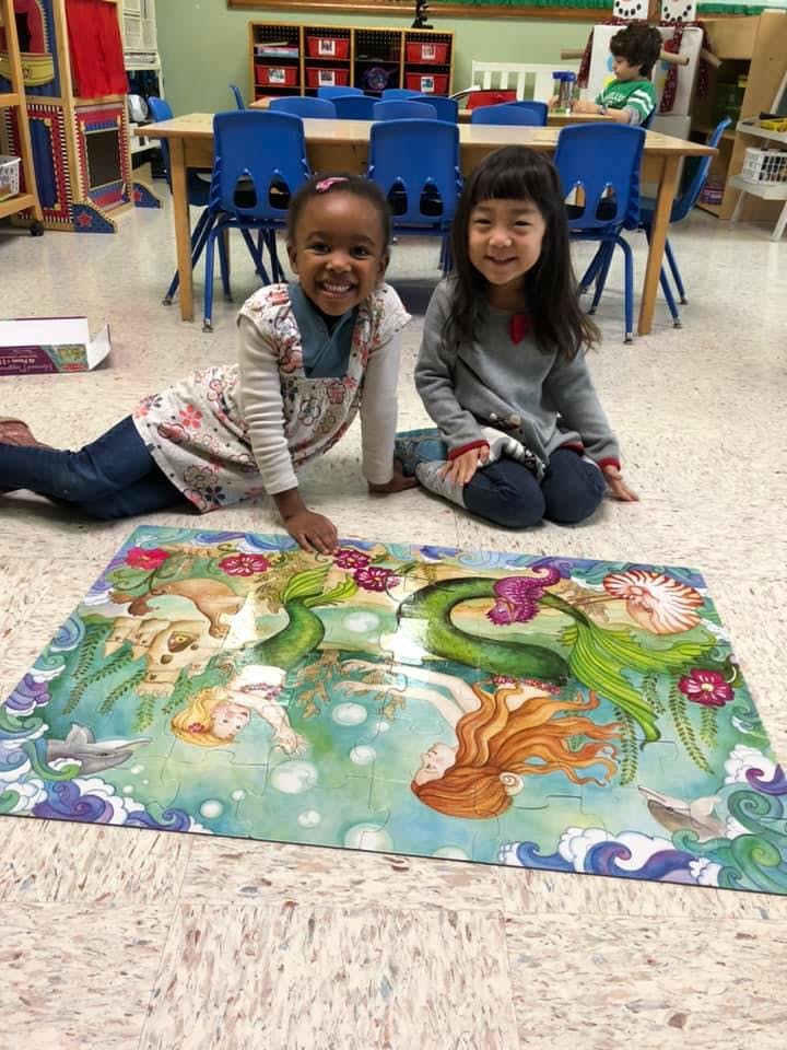 Two children on the floor next to a large mermaid puzzle in a classroom