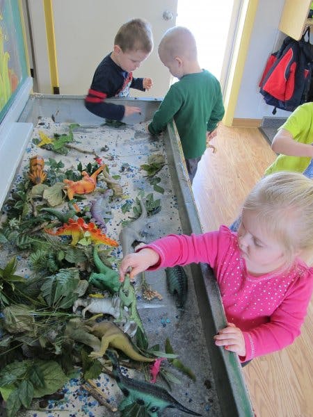 Three children playing with dinosaur toys in a sand table