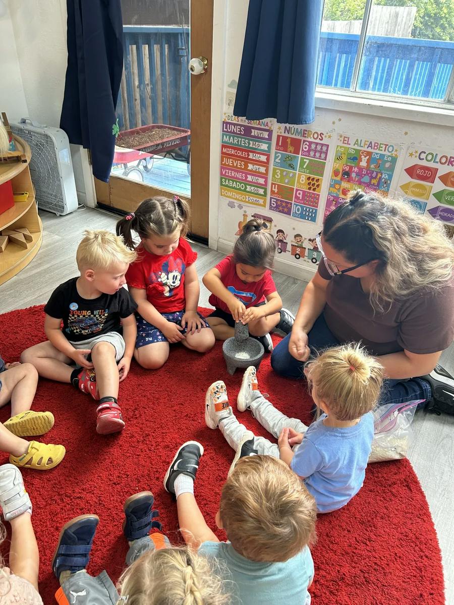 Group of children and an adult sitting on a rug using a mortar and pestle