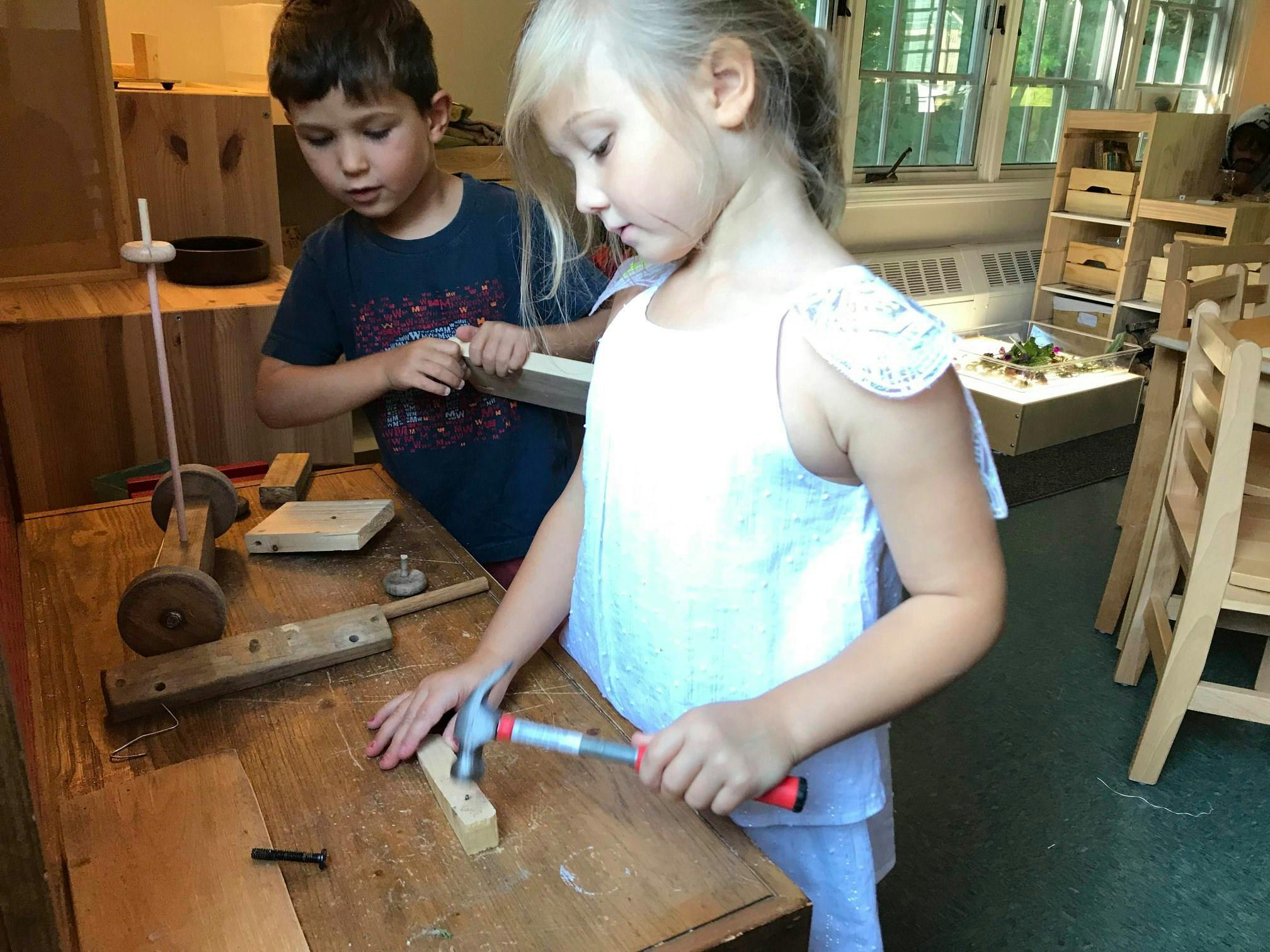 Child hammering a nail into wood, another child holding a piece of wood