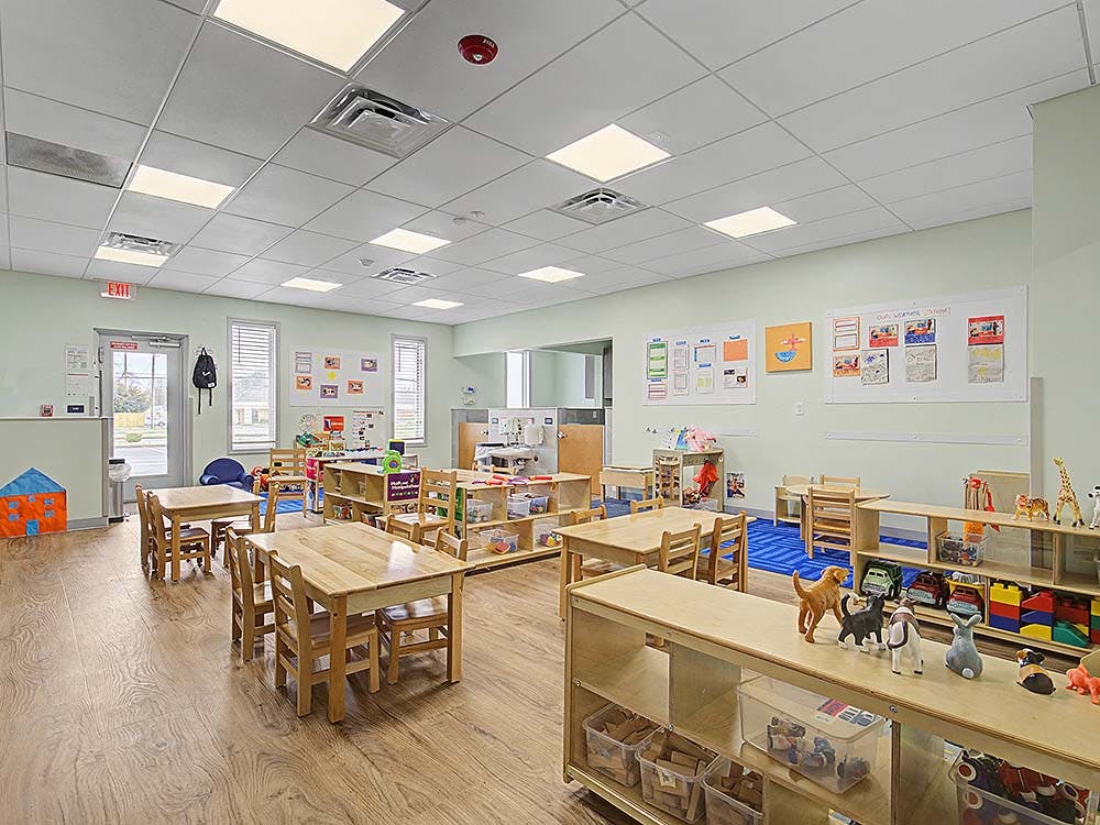Empty classroom with small wooden tables and chairs, shelves with toys