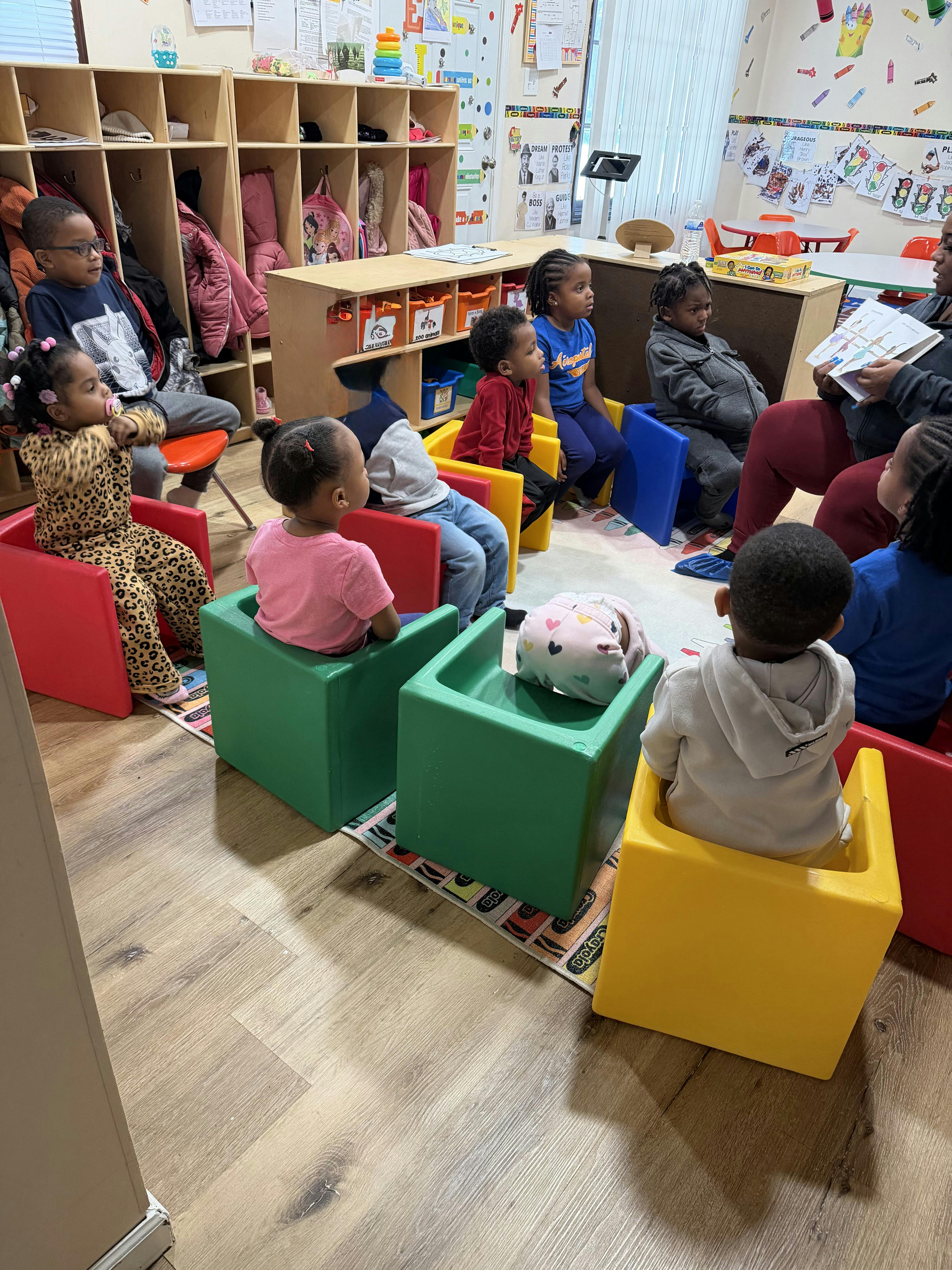 Group of children seated in a classroom, looking at an adult reading a book