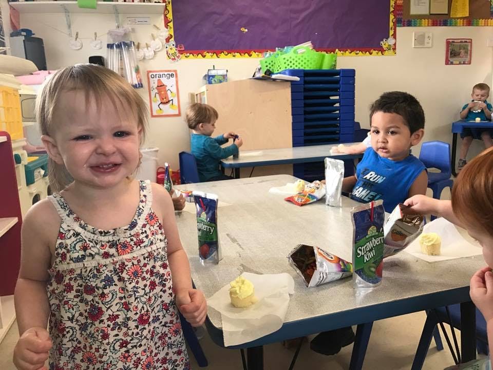 Child in floral top smiling at camera, other children at tables in classroom