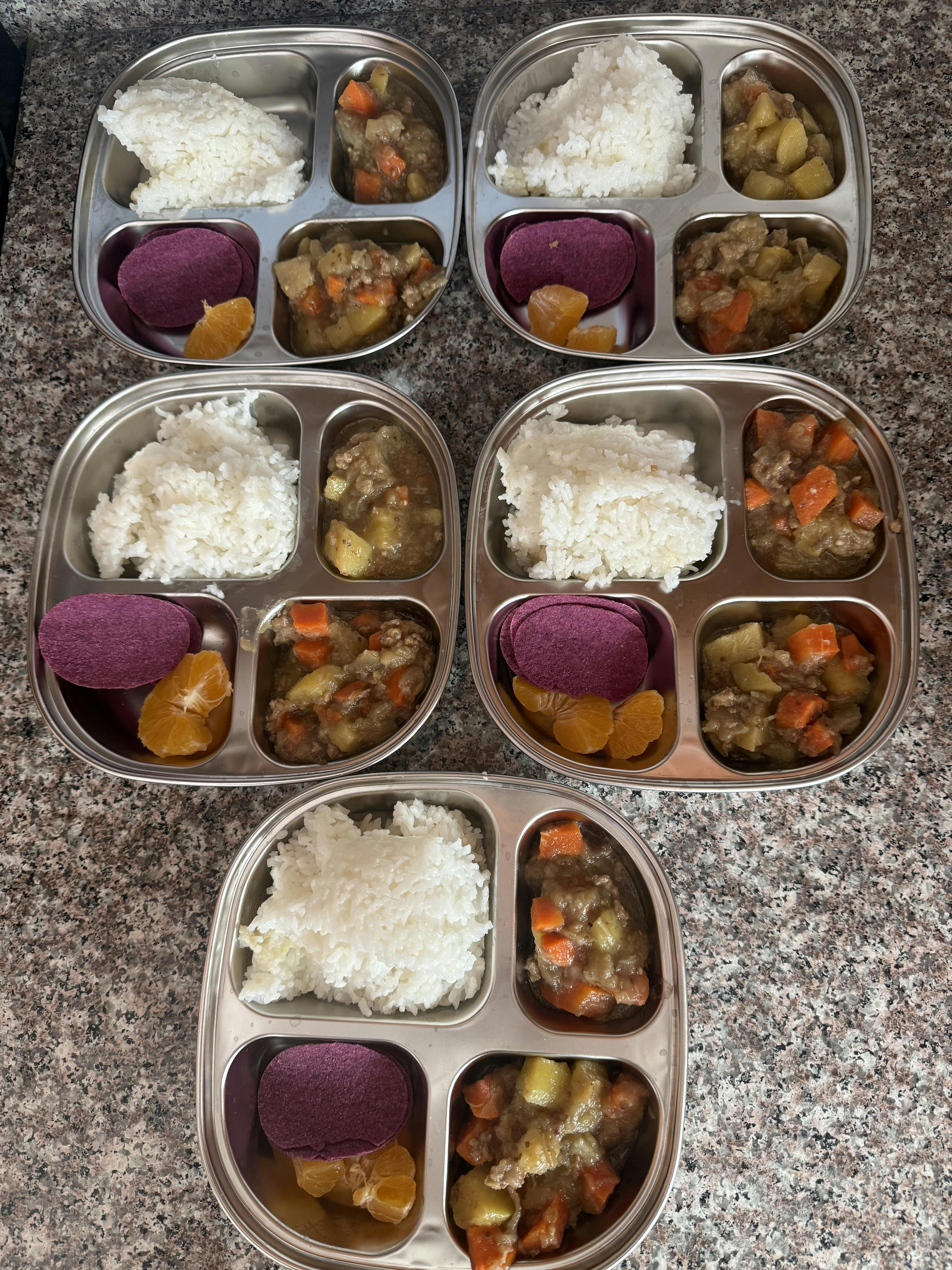 Five metal divided trays filled with rice, vegetable stew, purple disks, and fruit