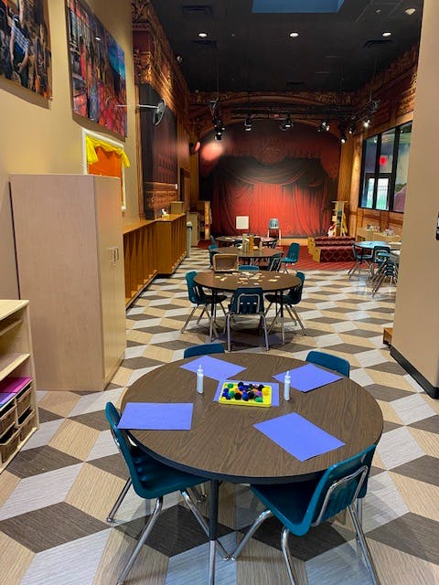 Classroom with small tables and chairs, a stage with red curtains in the background