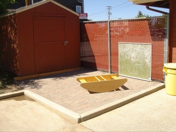 Wooden rocking boat on a paved area next to a red shed and fence