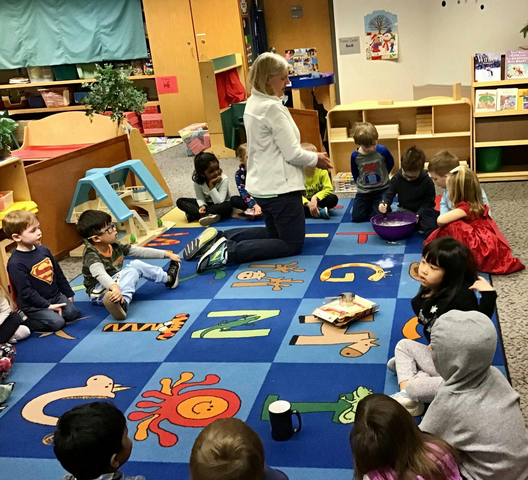 Adult kneeling on a rug with children sitting around in a classroom