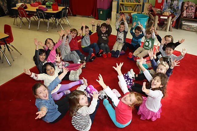 Group of children sitting in a circle on a red rug with hands raised