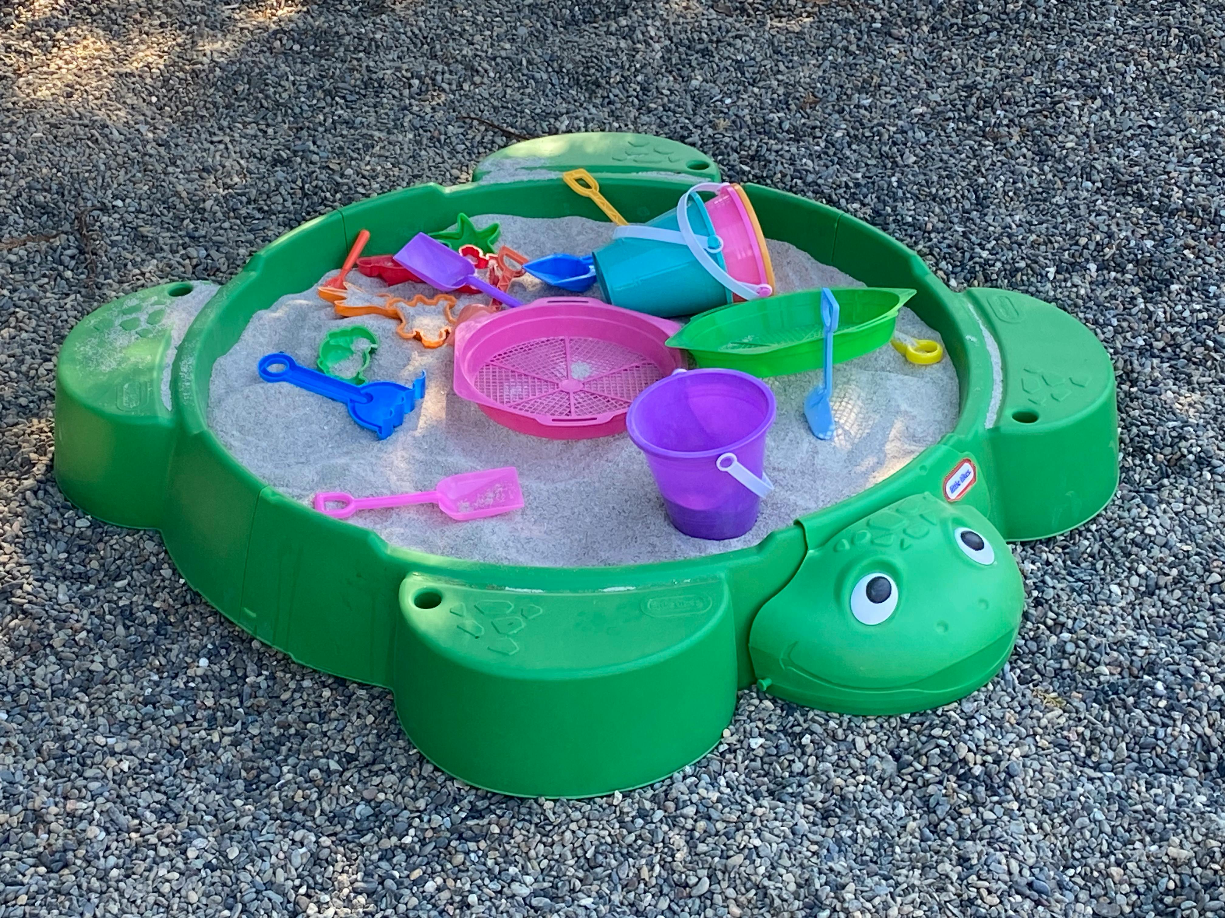 Green turtle sandbox filled with sand toys on a gravel surface