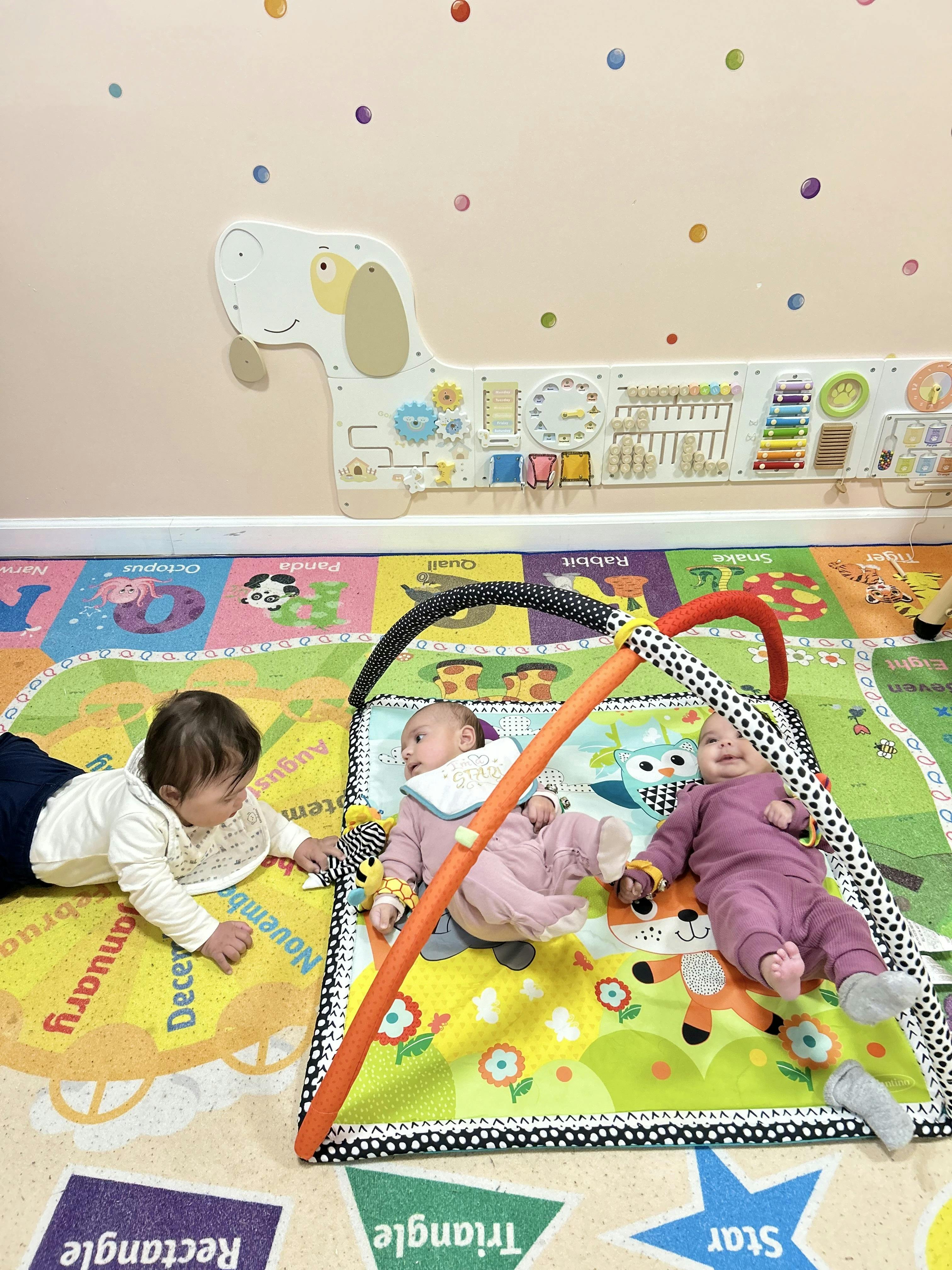 Three children playing on a colorful mat with a dog-shaped activity board on the wall