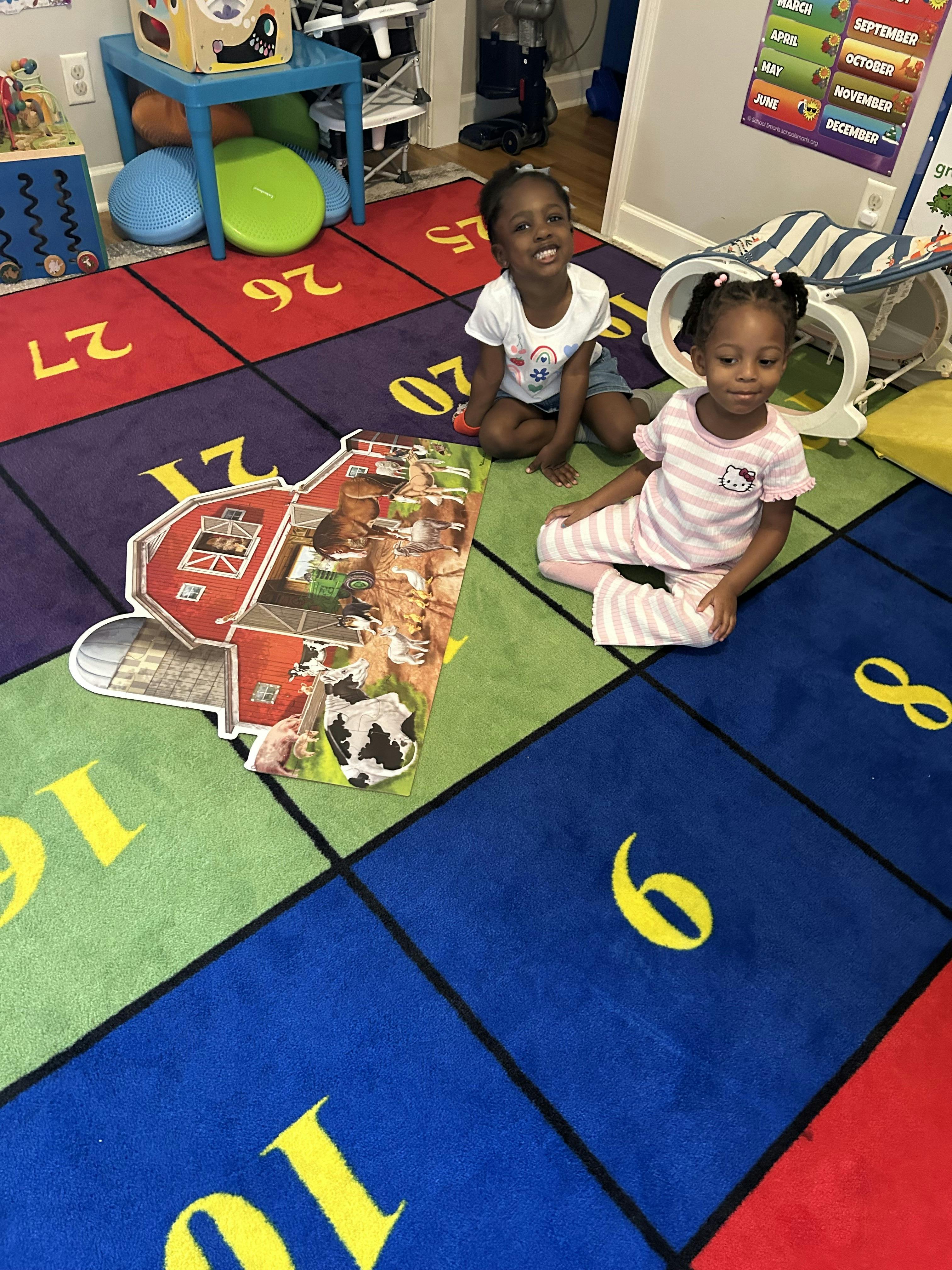 Two children sitting on a numbered rug next to a barn-themed floor puzzle