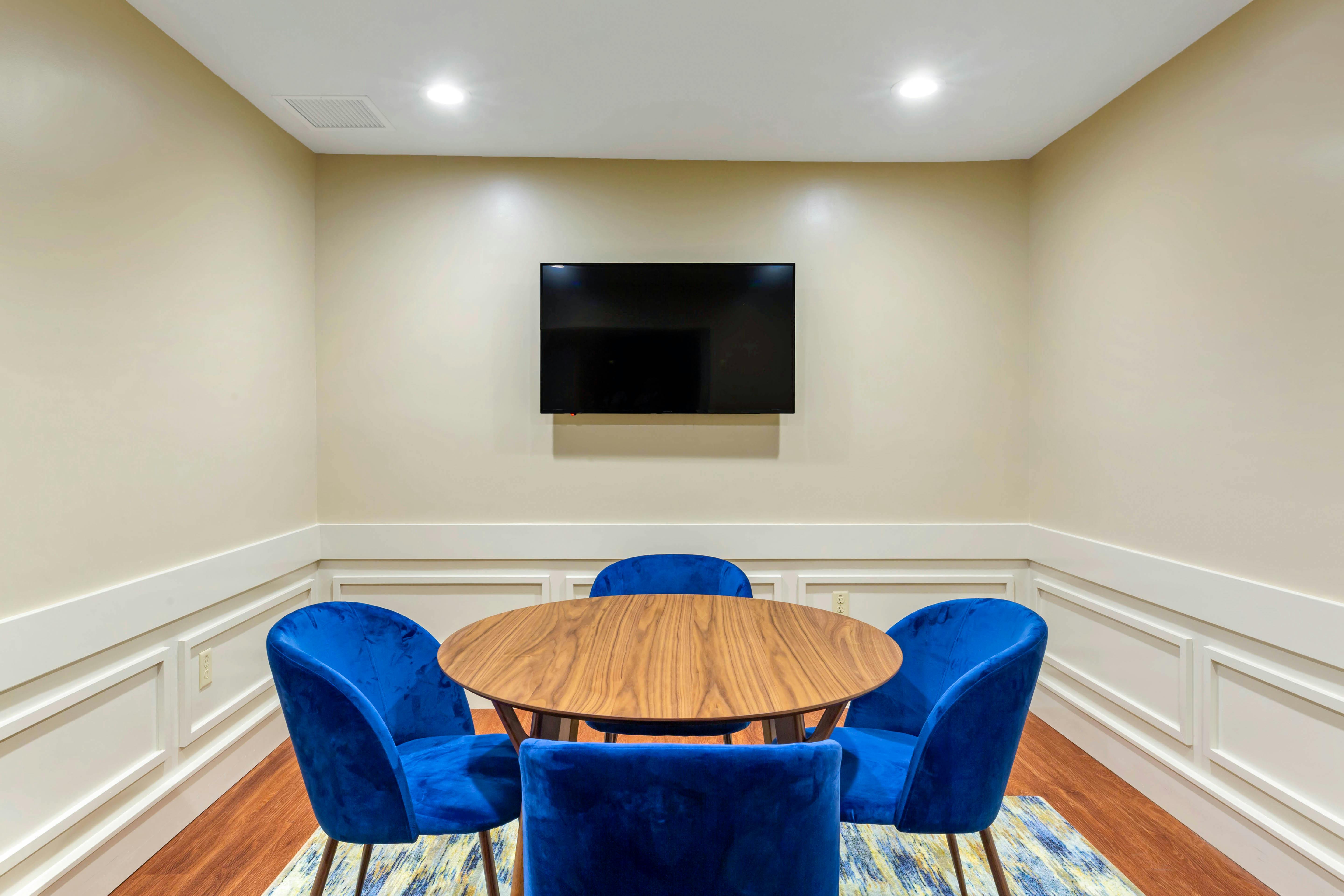 Round wooden table surrounded by four blue chairs with a TV mounted on the wall