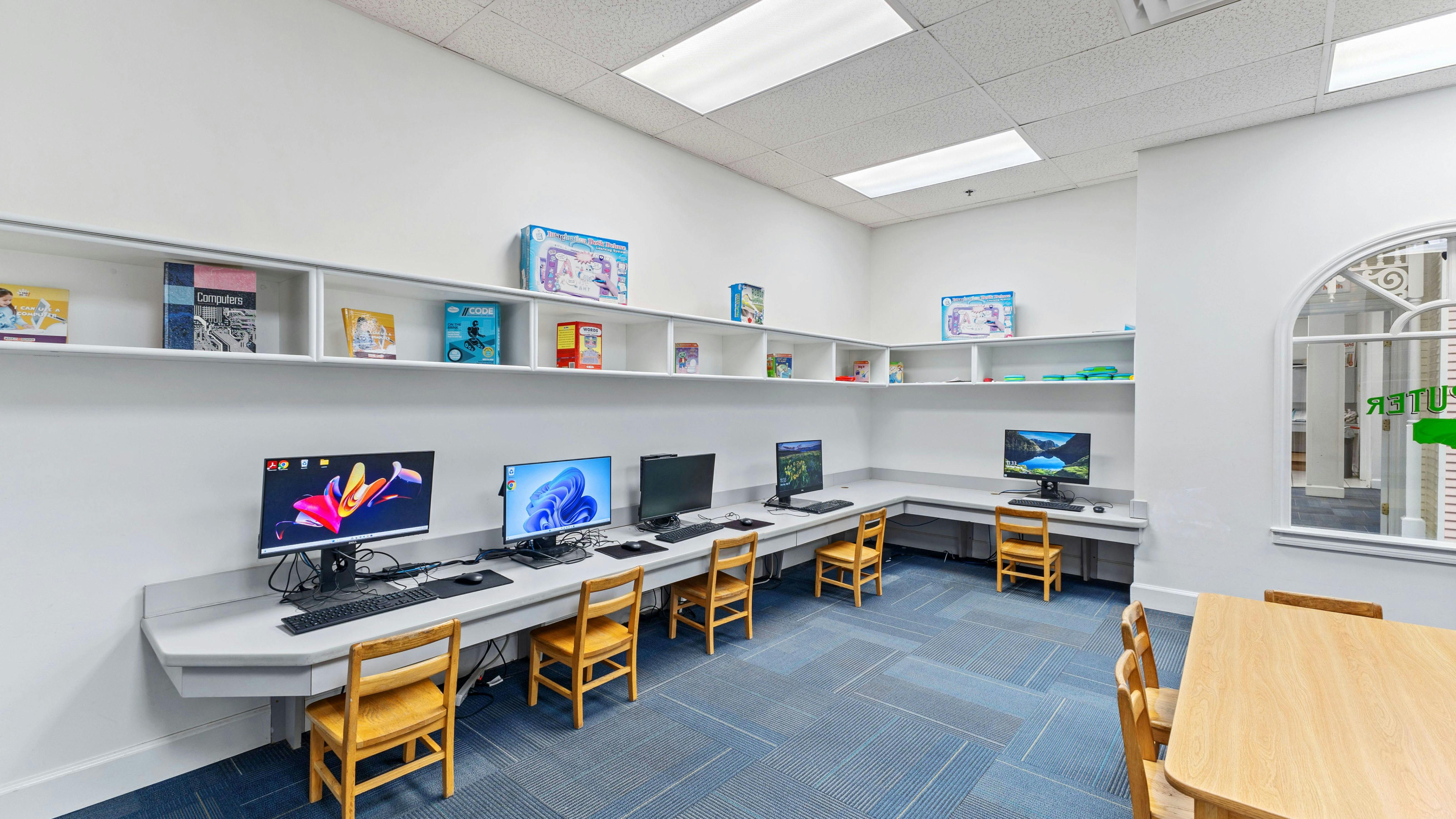 A computer lab with several desktop computers and small chairs along a white wall