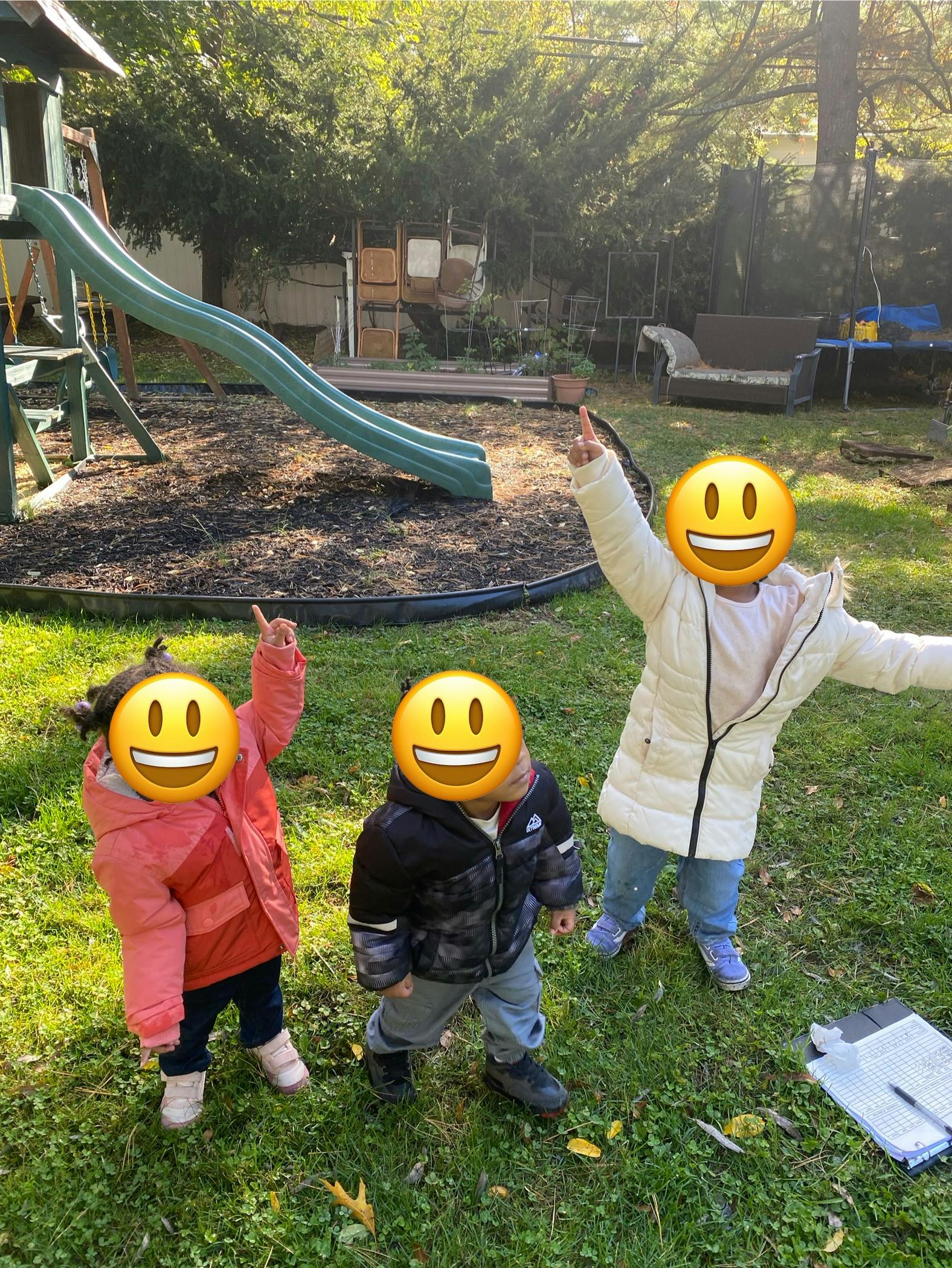 Three children in winter coats pointing up in a grassy yard