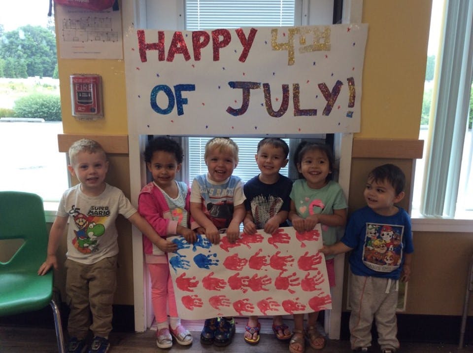Five children holding a banner with handprints, under a "Happy 4th of July!" sign