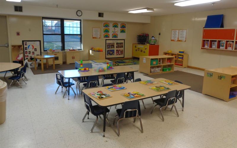 Empty classroom with small tables and chairs, toys on shelves, and windows