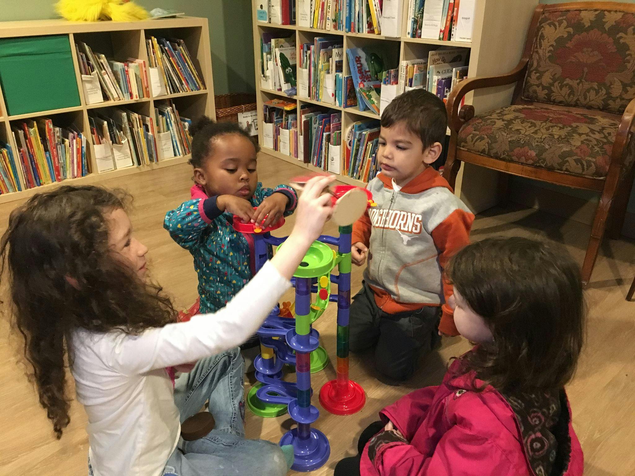 Four children playing with a colorful toy in a room with bookshelves