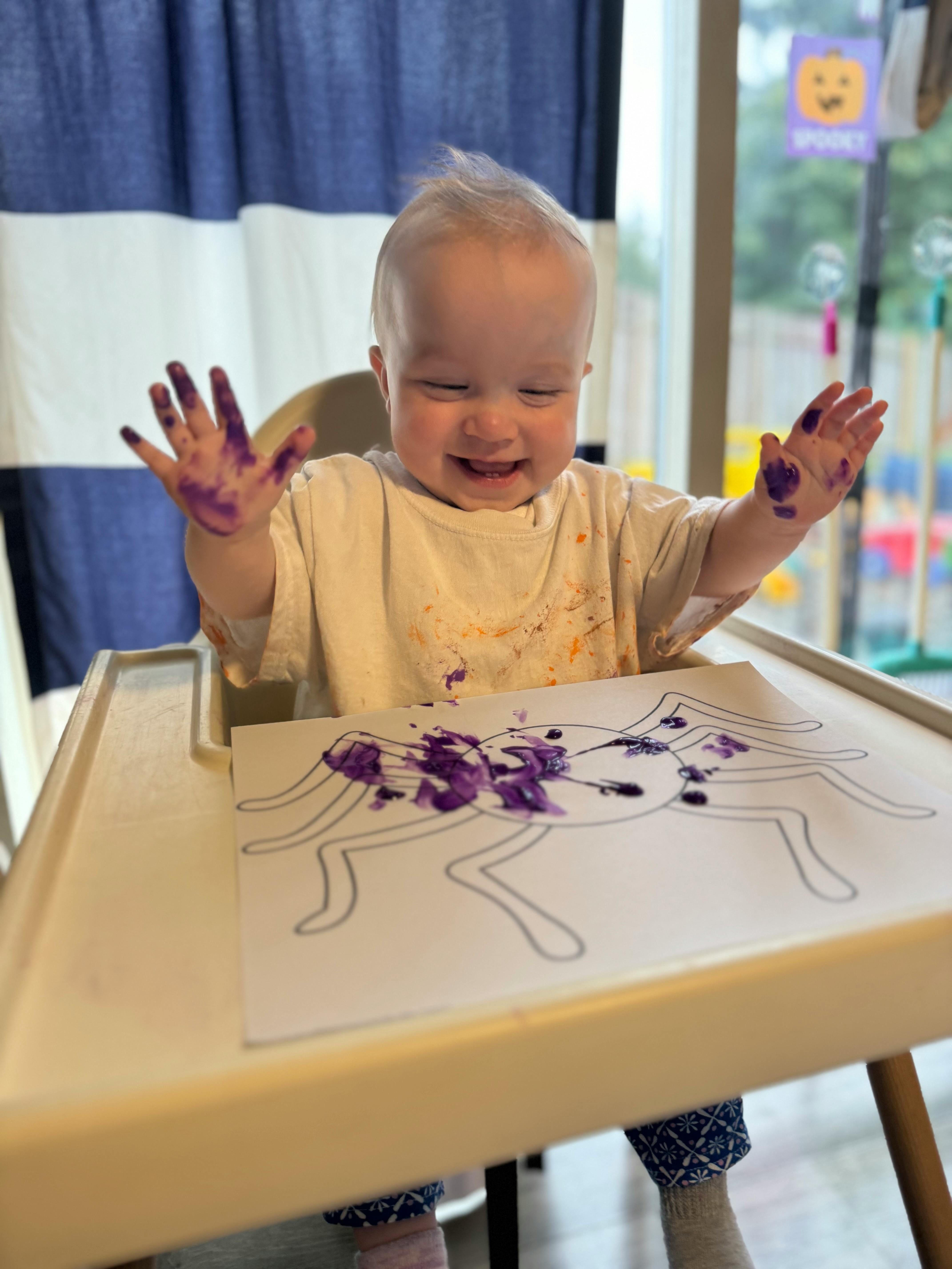 Child in high chair with purple paint on hands and paper