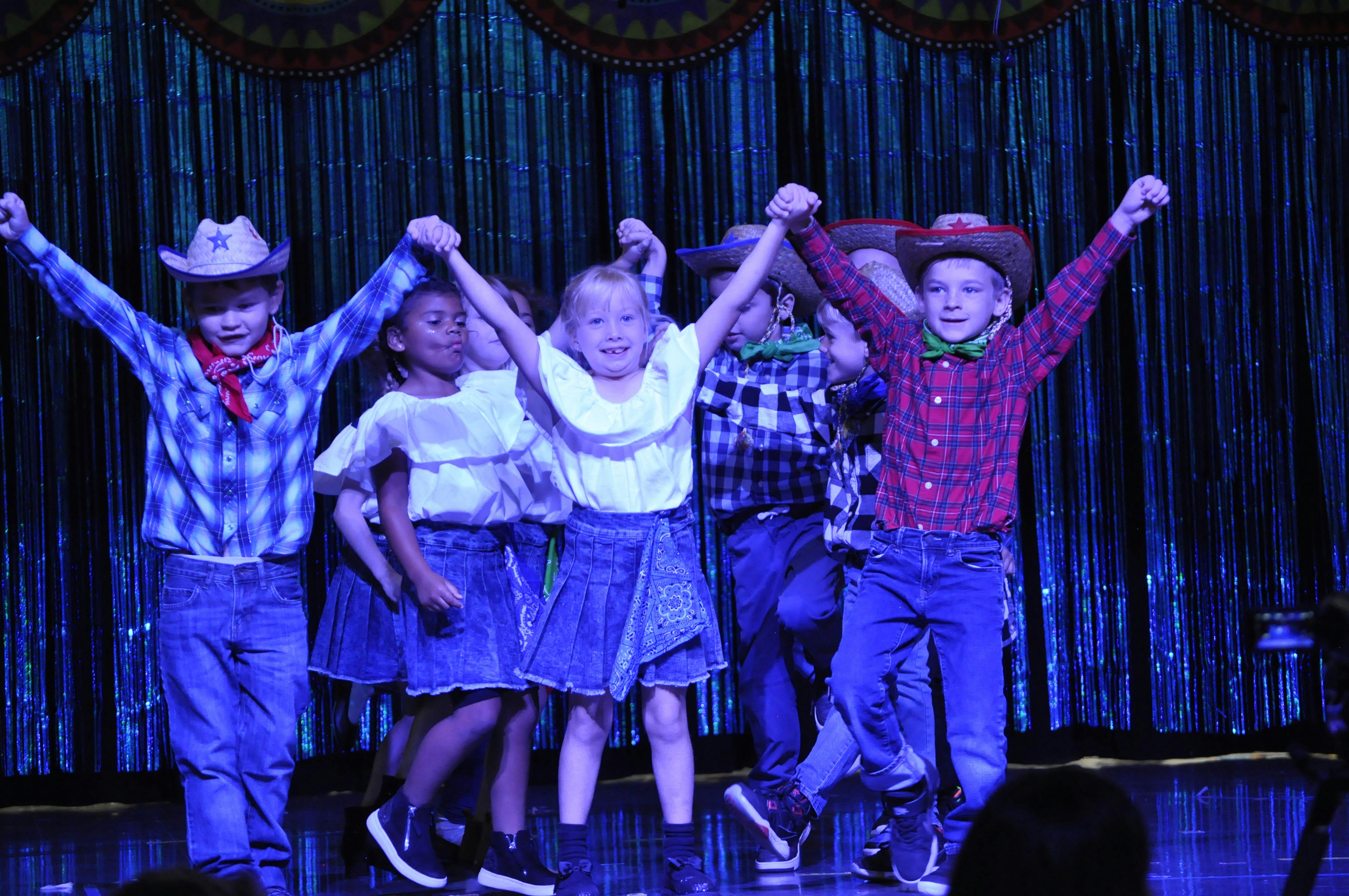 Children in cowboy hats and skirts or jeans holding hands and raising arms