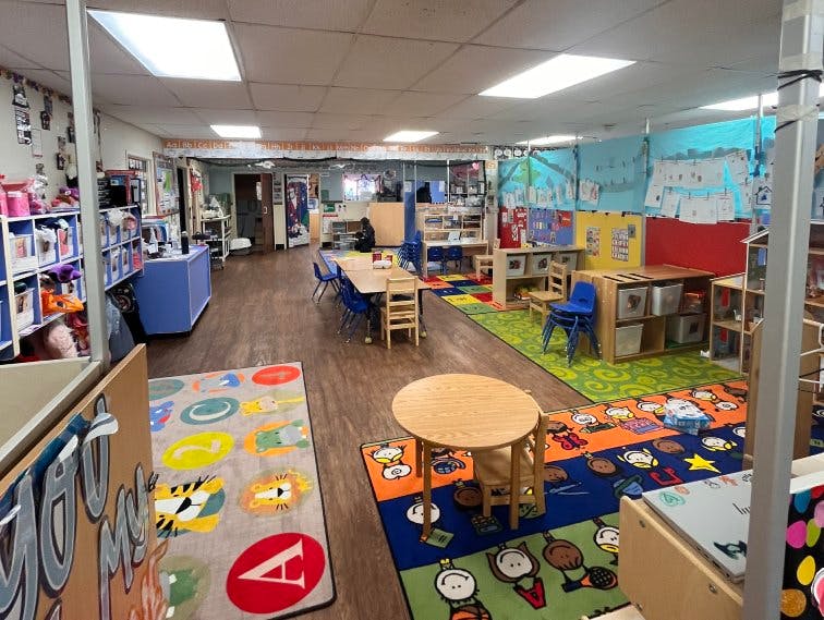 A classroom with small tables and chairs, colorful rugs, and learning materials