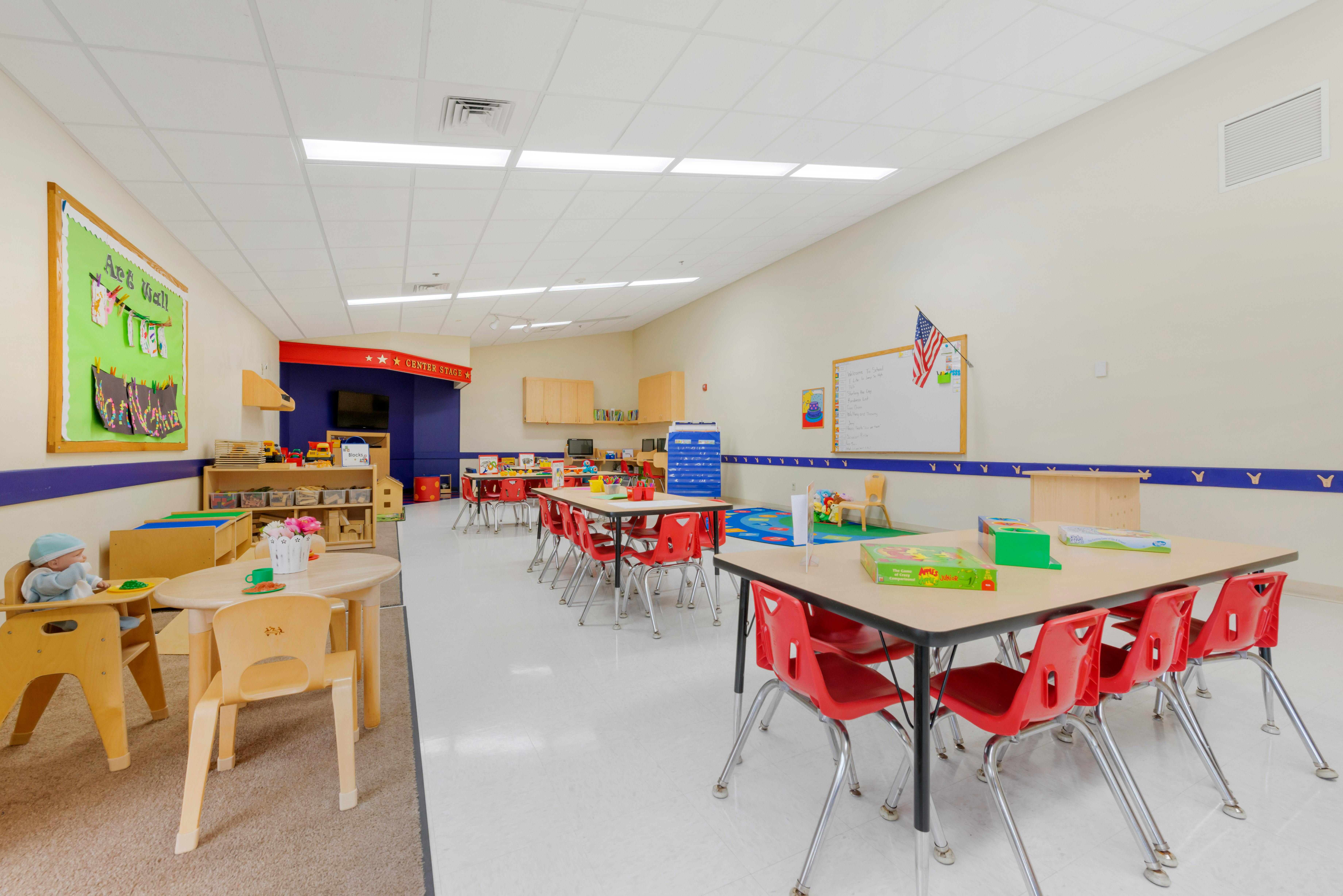 A colorful classroom with tables and red chairs, an art wall, and a play area