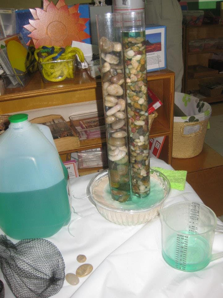Two clear tubes filled with rocks and water in a bowl, next to a jug of liquid