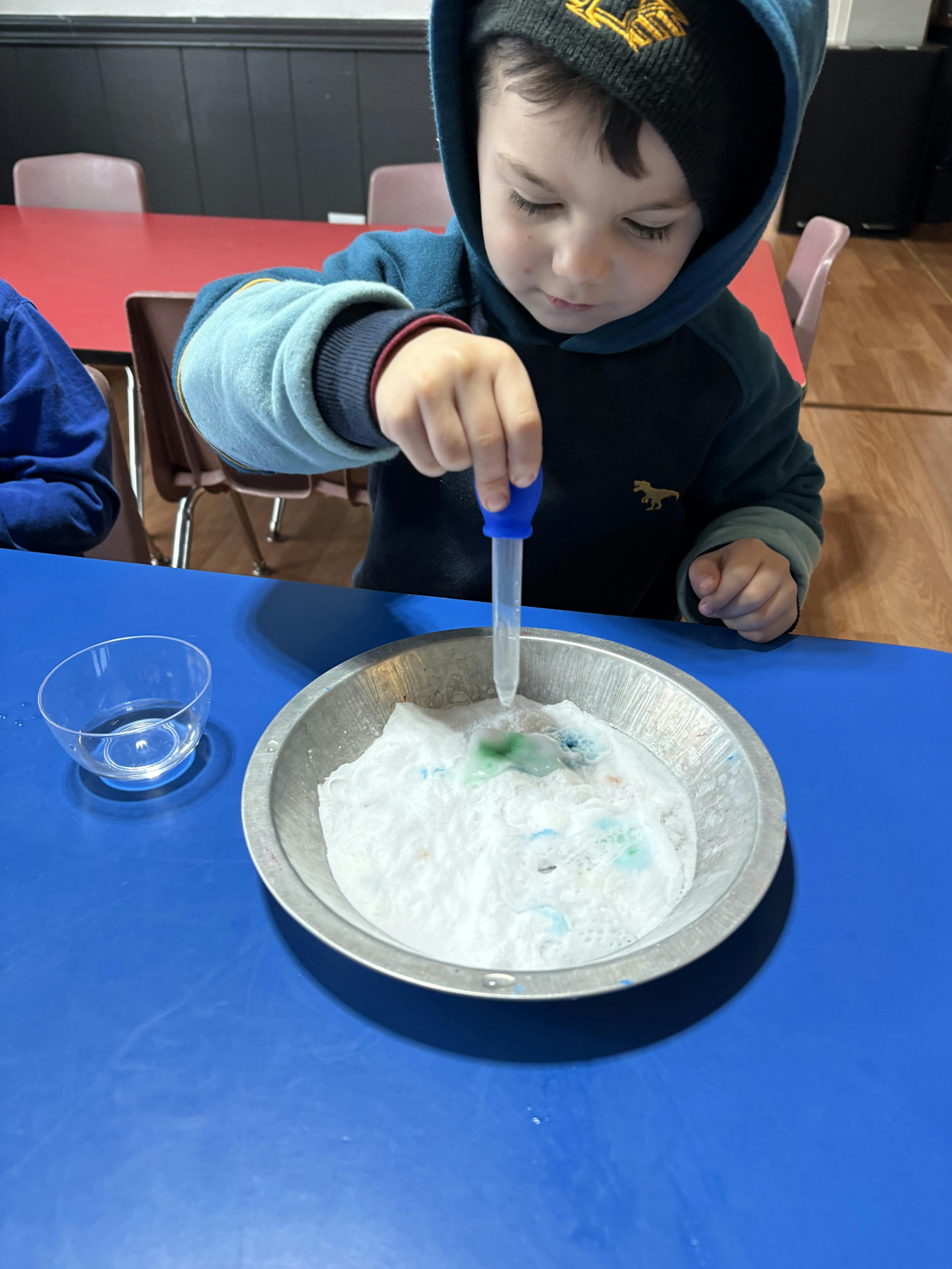 Child using a dropper to add liquid to a white foamy substance in a metal dish