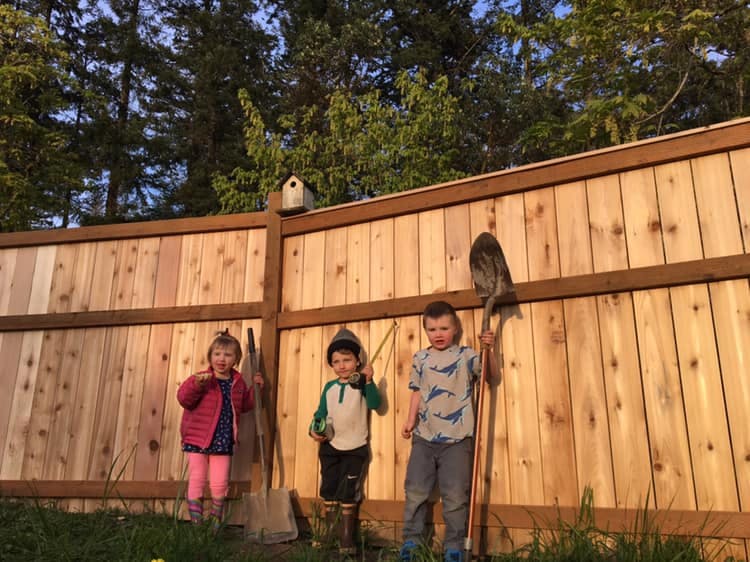 Three children standing in front of a wooden fence with gardening tools
