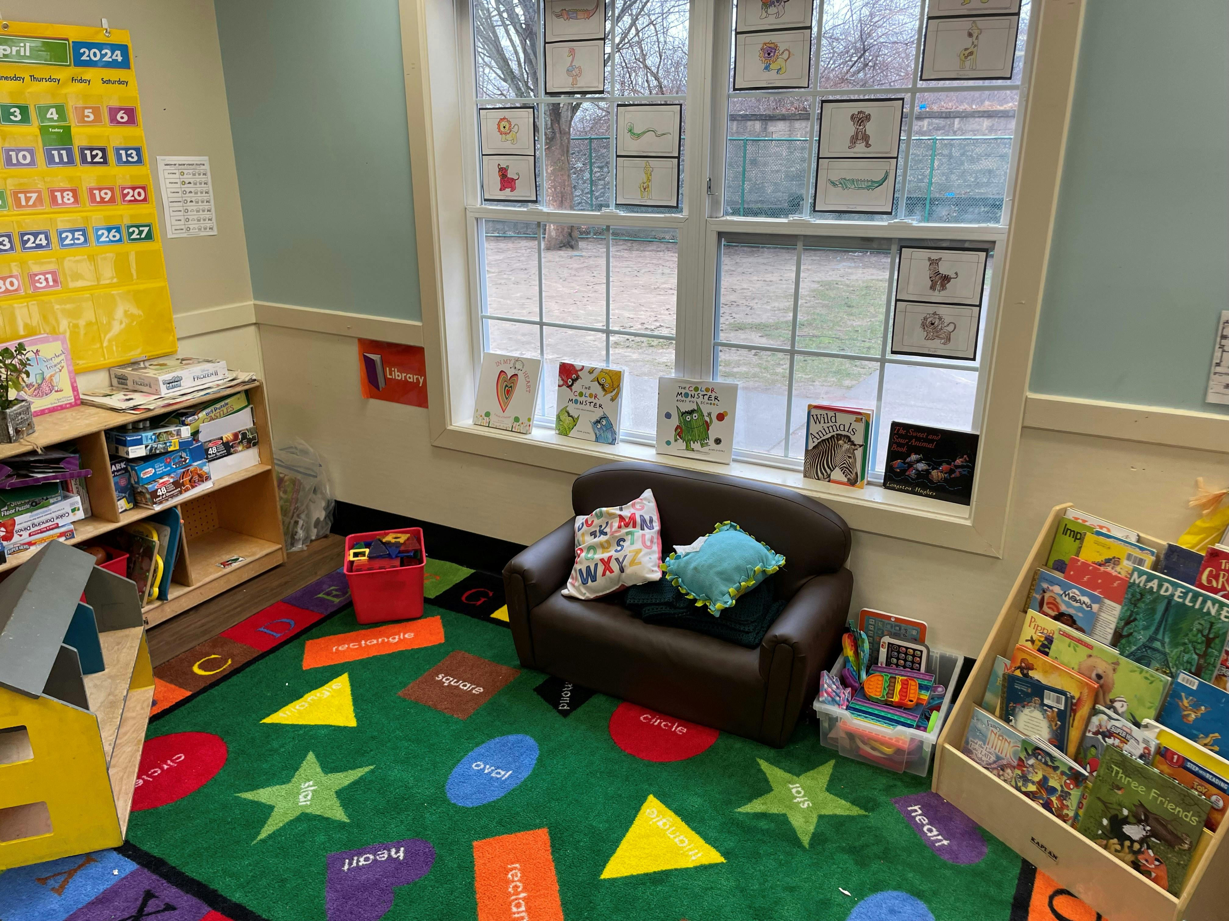 Classroom corner with an armchair, colorful rug with shapes, bookshelves, and window