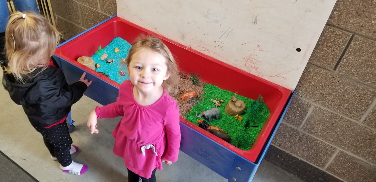 Child standing next to a sensory bin with another child in the background
