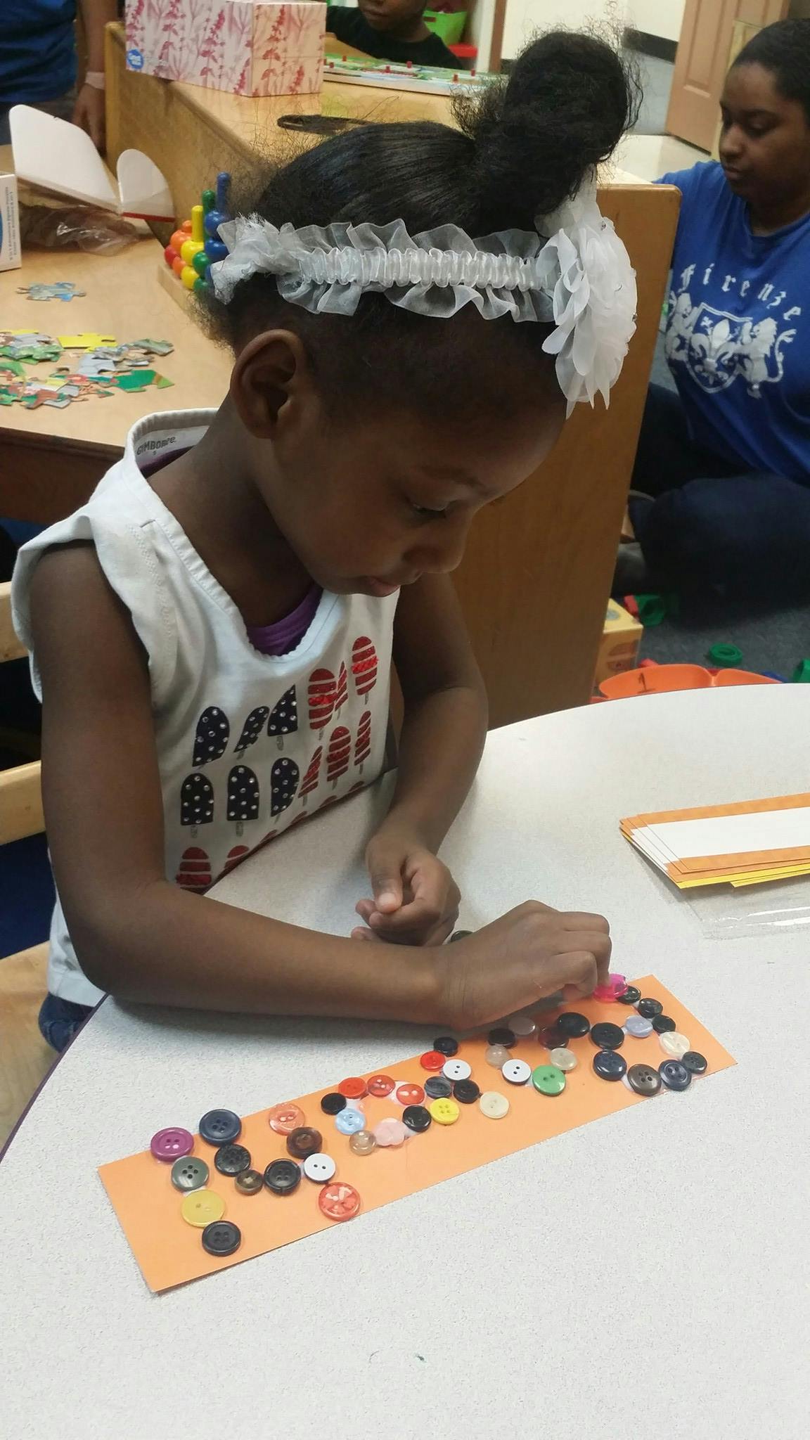 Child placing buttons on an orange paper strip at a table