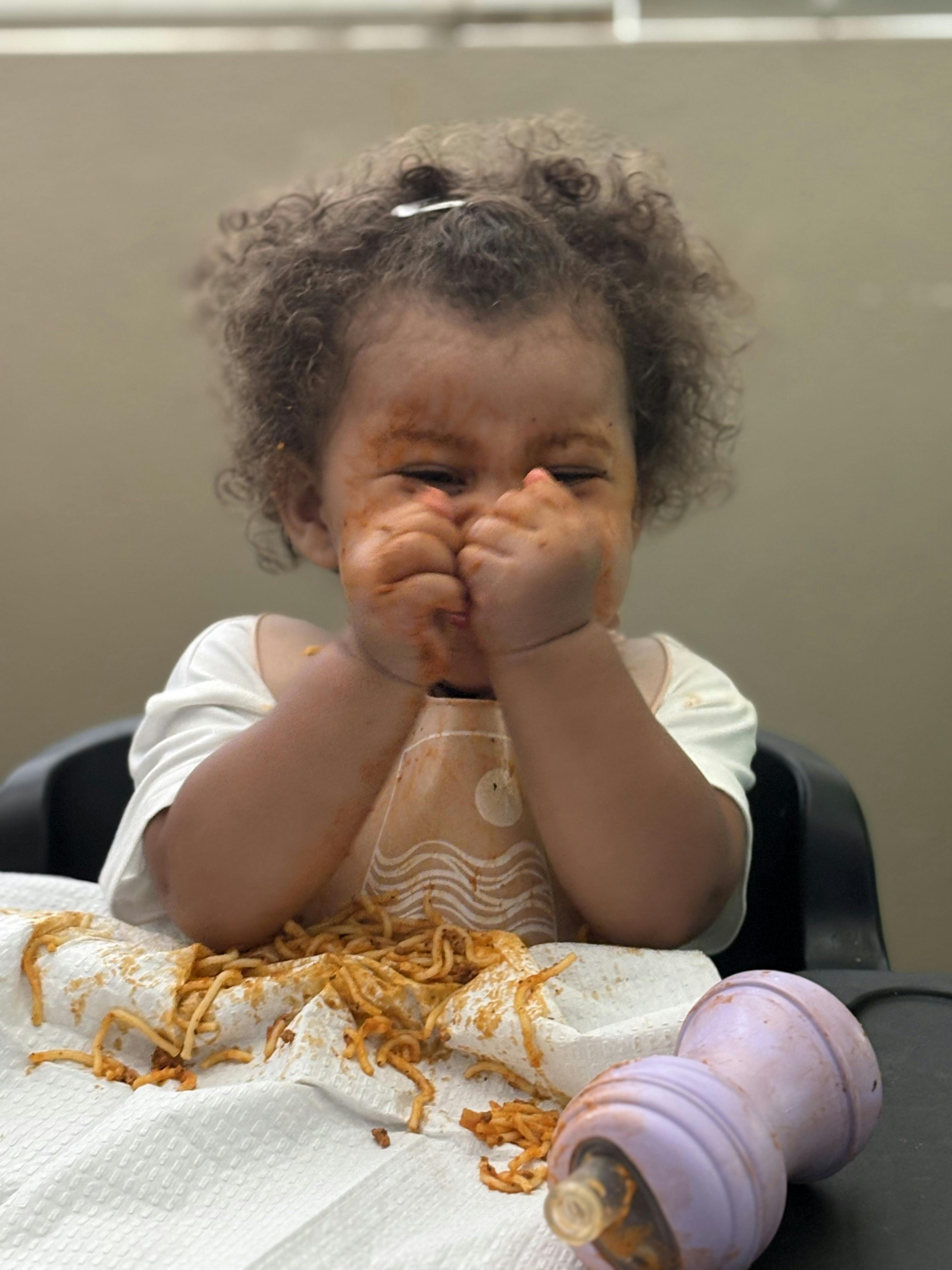 Child in highchair with messy hands and face from eating pasta