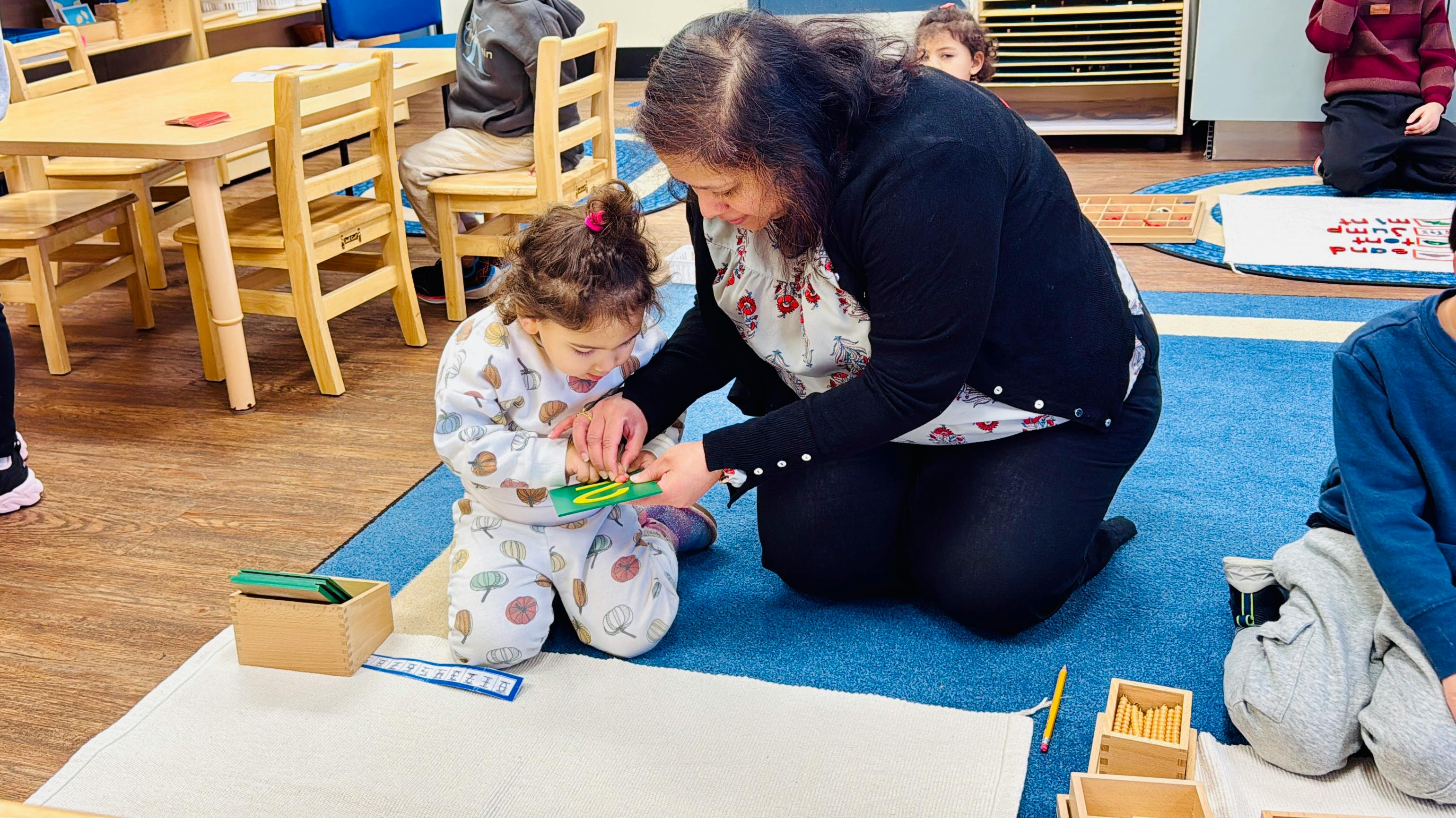 Adult and child on floor, engaged with learning materials in a classroom