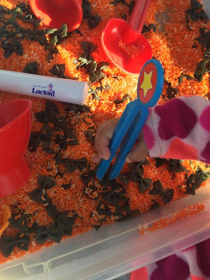 Child's hand using blue tongs in a bin of orange rice and green pasta