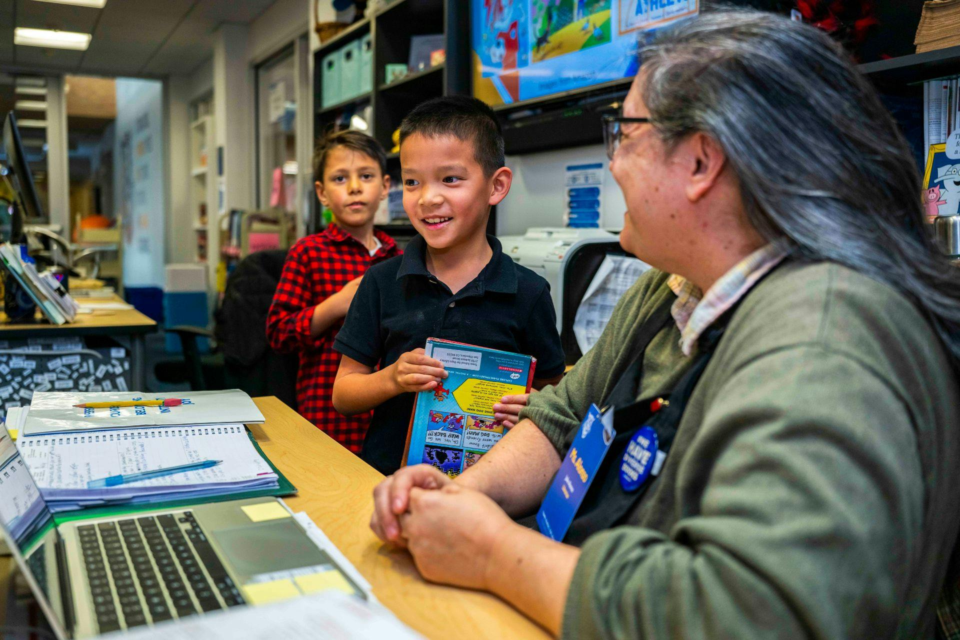 two children standing by a desk with an adult, one child holds a book