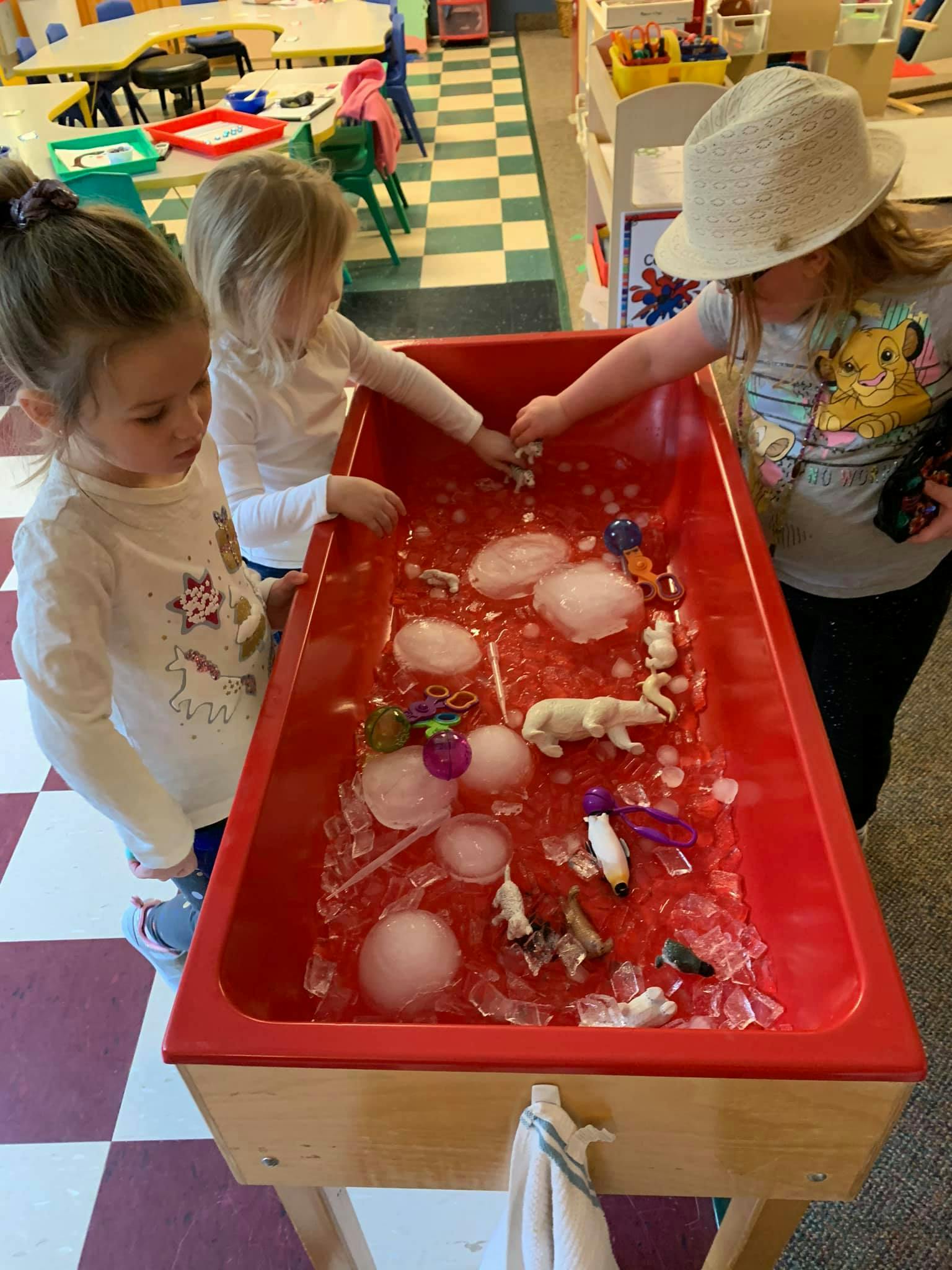 Three children playing with ice and animal figures in a red sensory bin