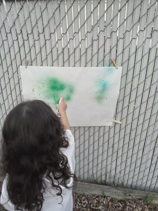 Child spraying paint onto a piece of paper attached to a chain link fence