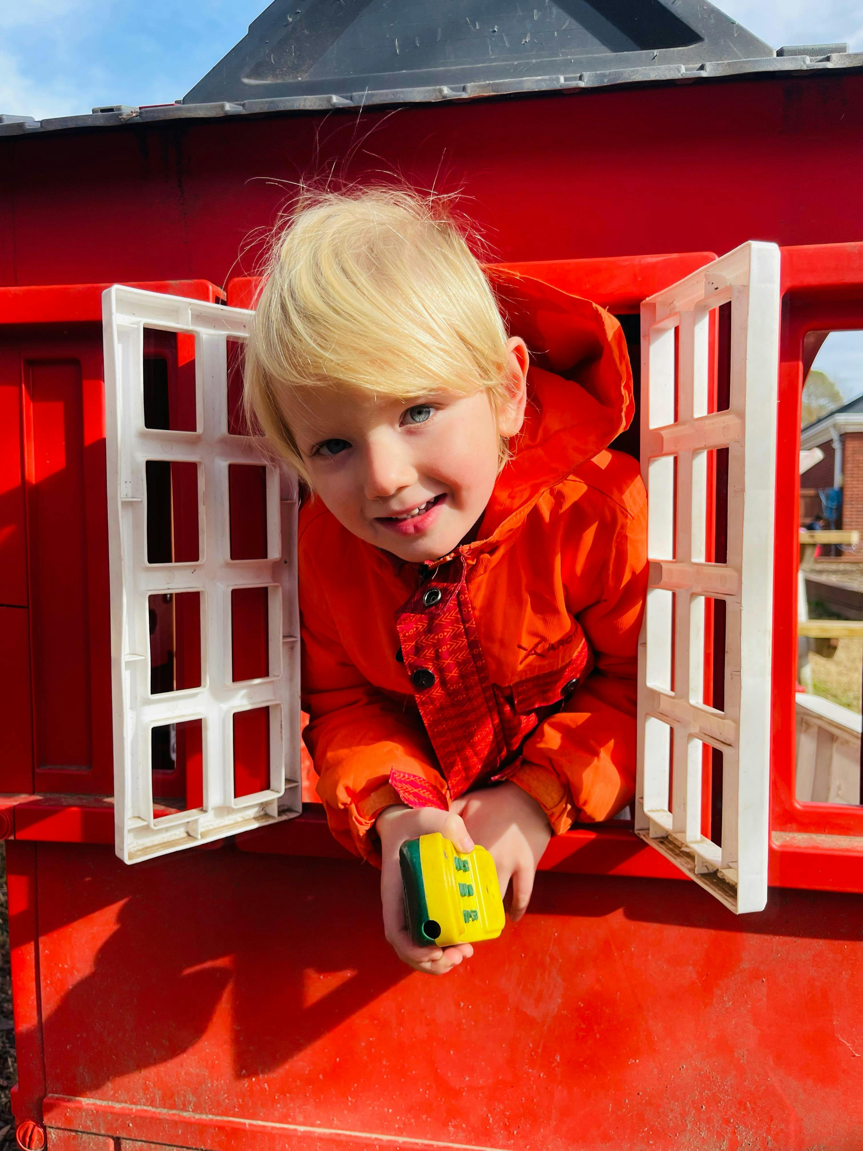 Child looking out a red playhouse window with a yellow toy