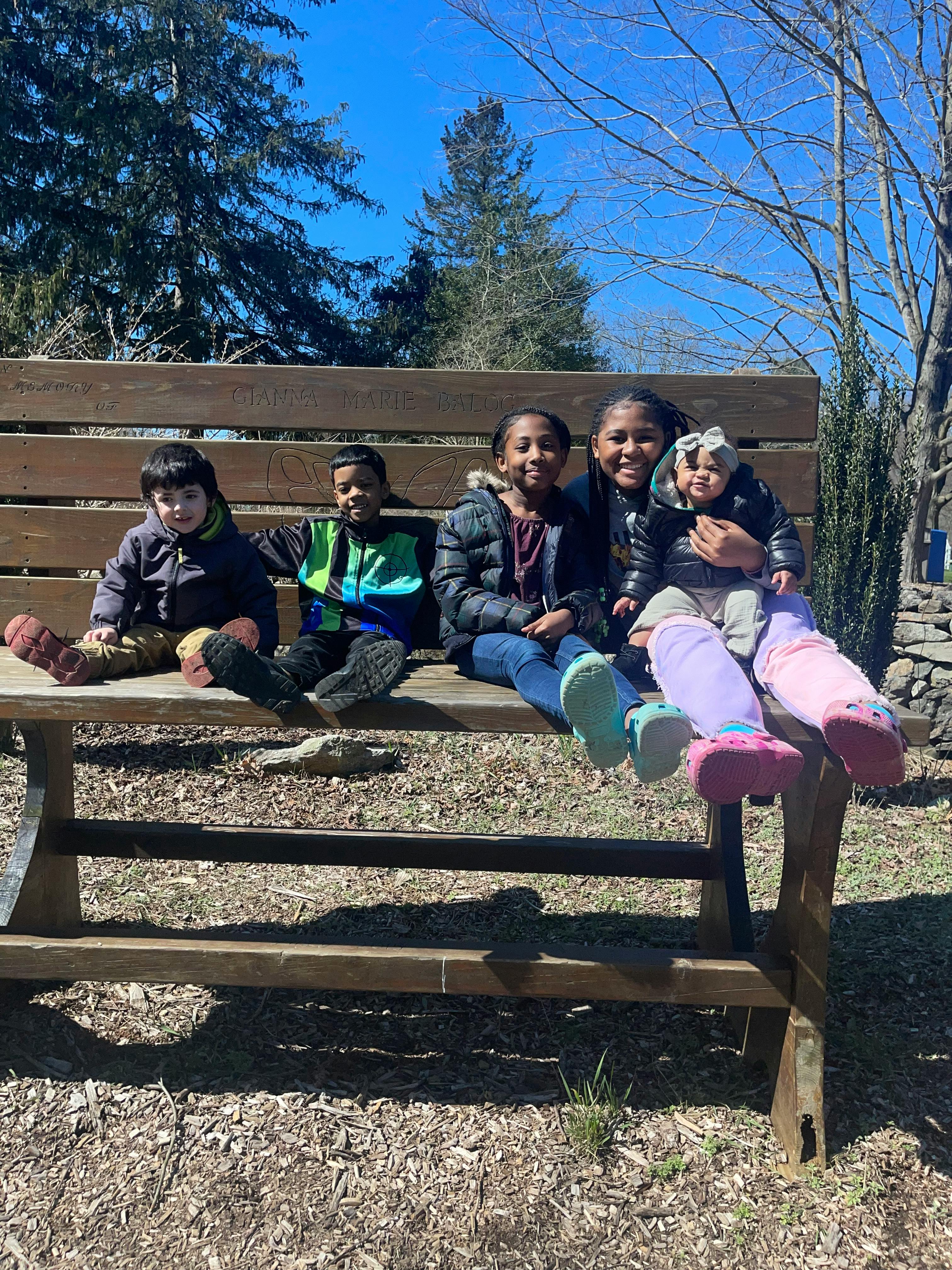 Five children sitting on a wooden picnic table bench outdoors