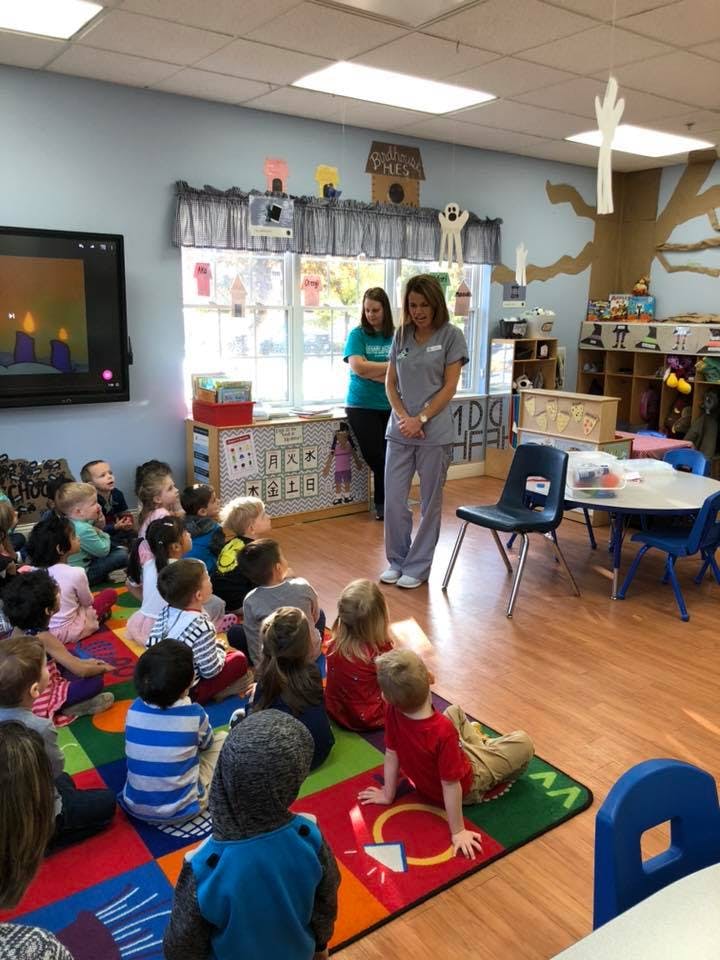 Two people standing in a classroom with children sitting on a colorful rug