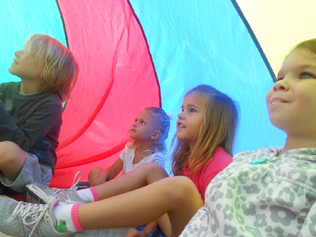 Four children sitting under a colorful parachute looking up