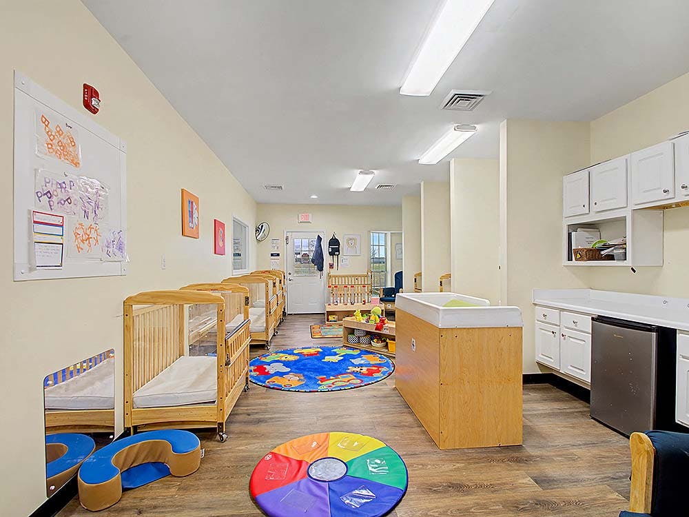 A brightly lit nursery room with several wooden cribs and playmats on the floor