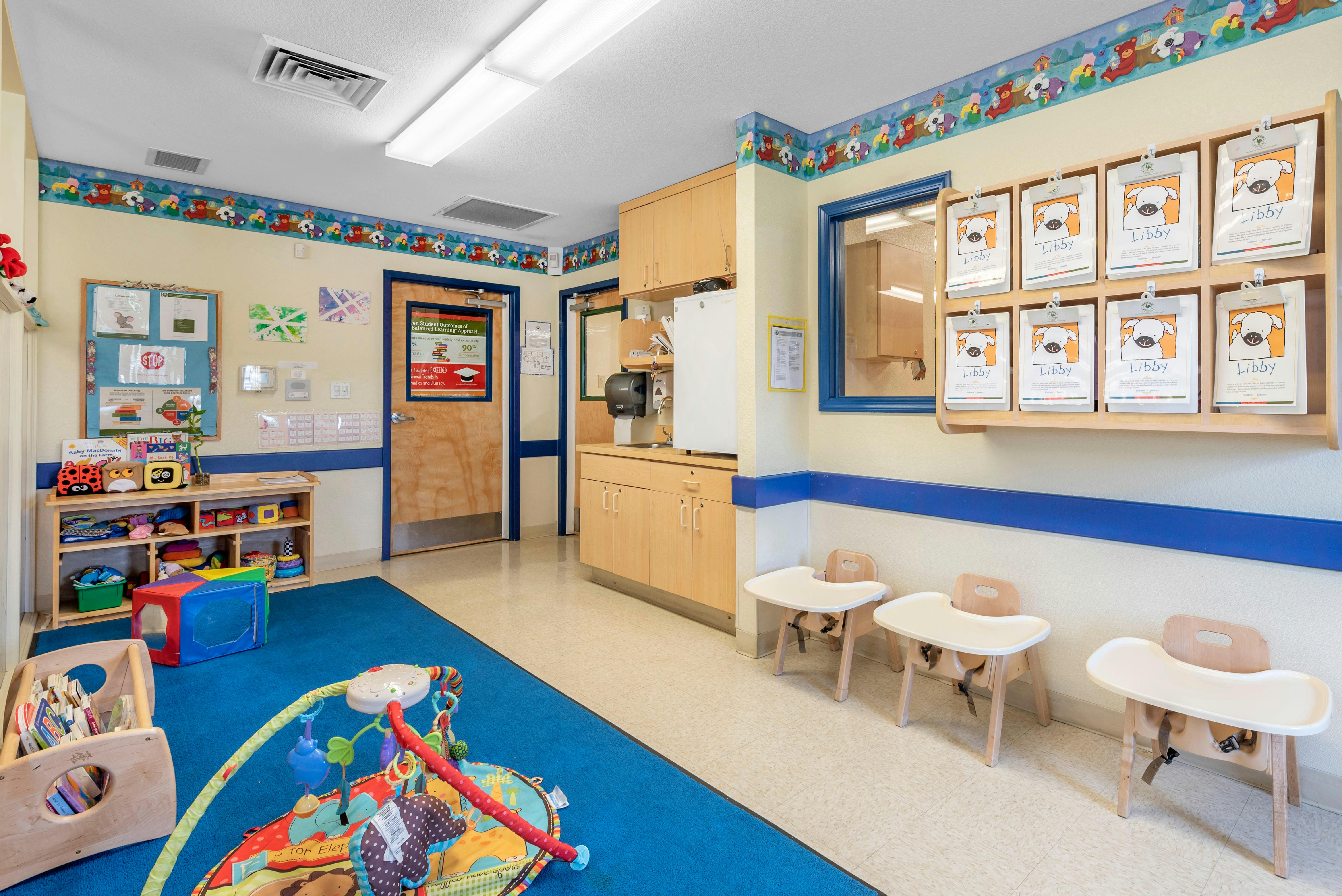A brightly lit classroom with a blue rug, small tables, and storage shelves