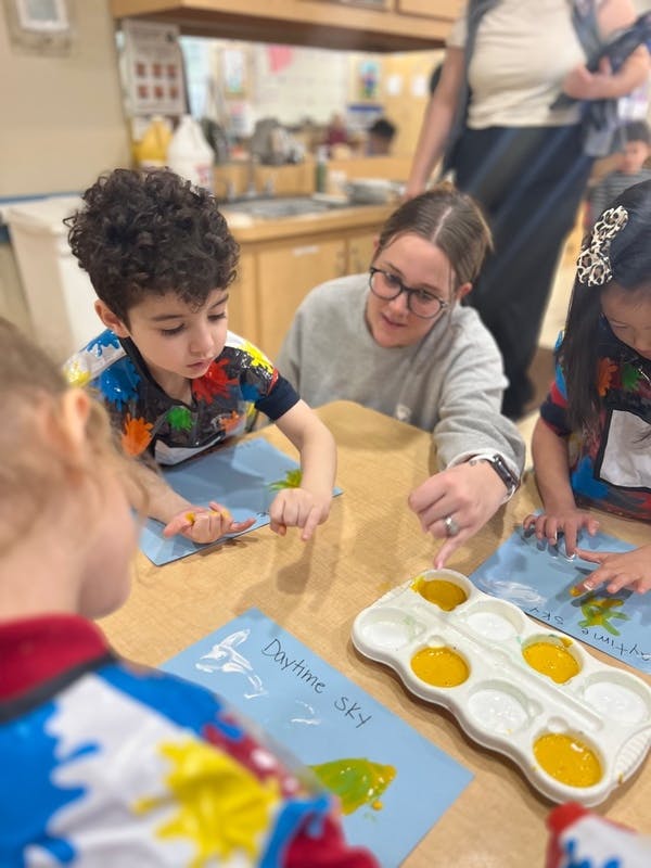 Adult helping children paint on blue paper at a table