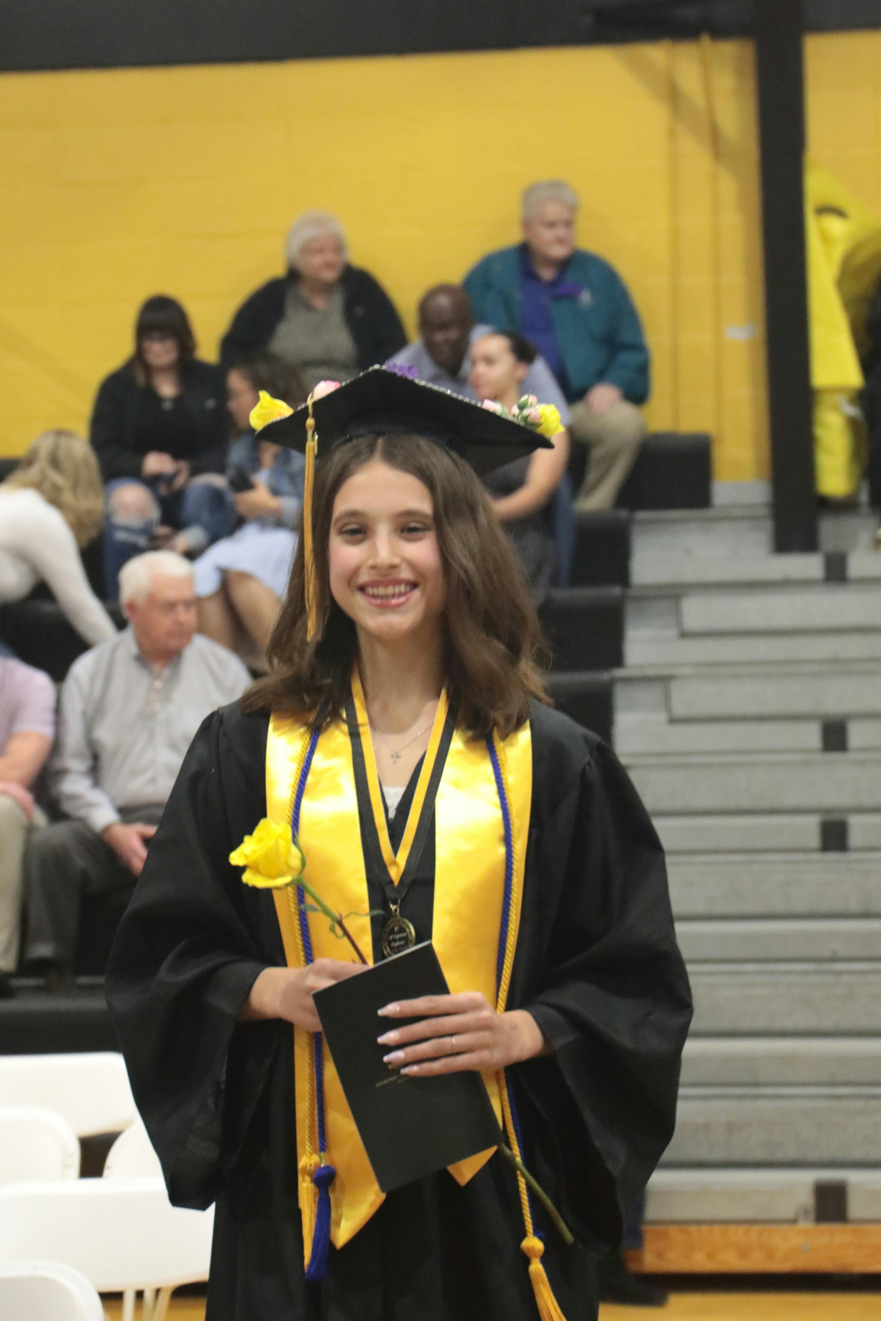 A teen wearing a graduation cap and gown holding a yellow rose and diploma