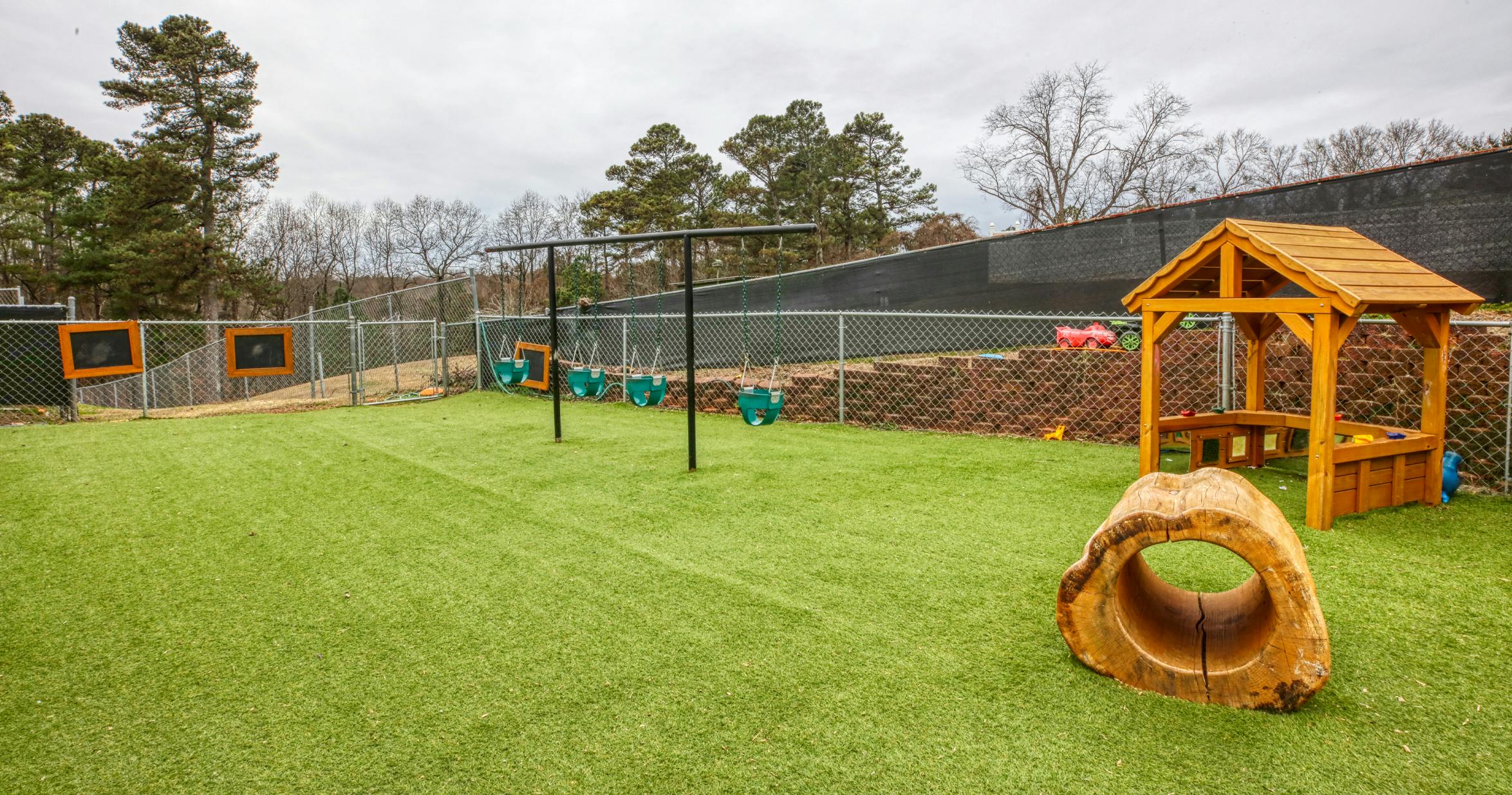 A fenced playground with artificial turf, swings, a wooden playhouse, and a log tunnel
