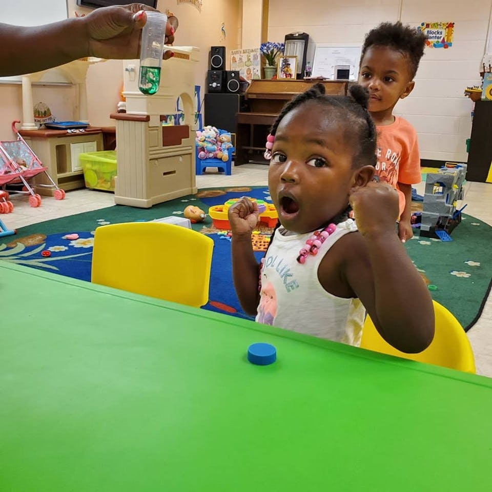 Child with open mouth and clenched fists looking at a person holding a bottle