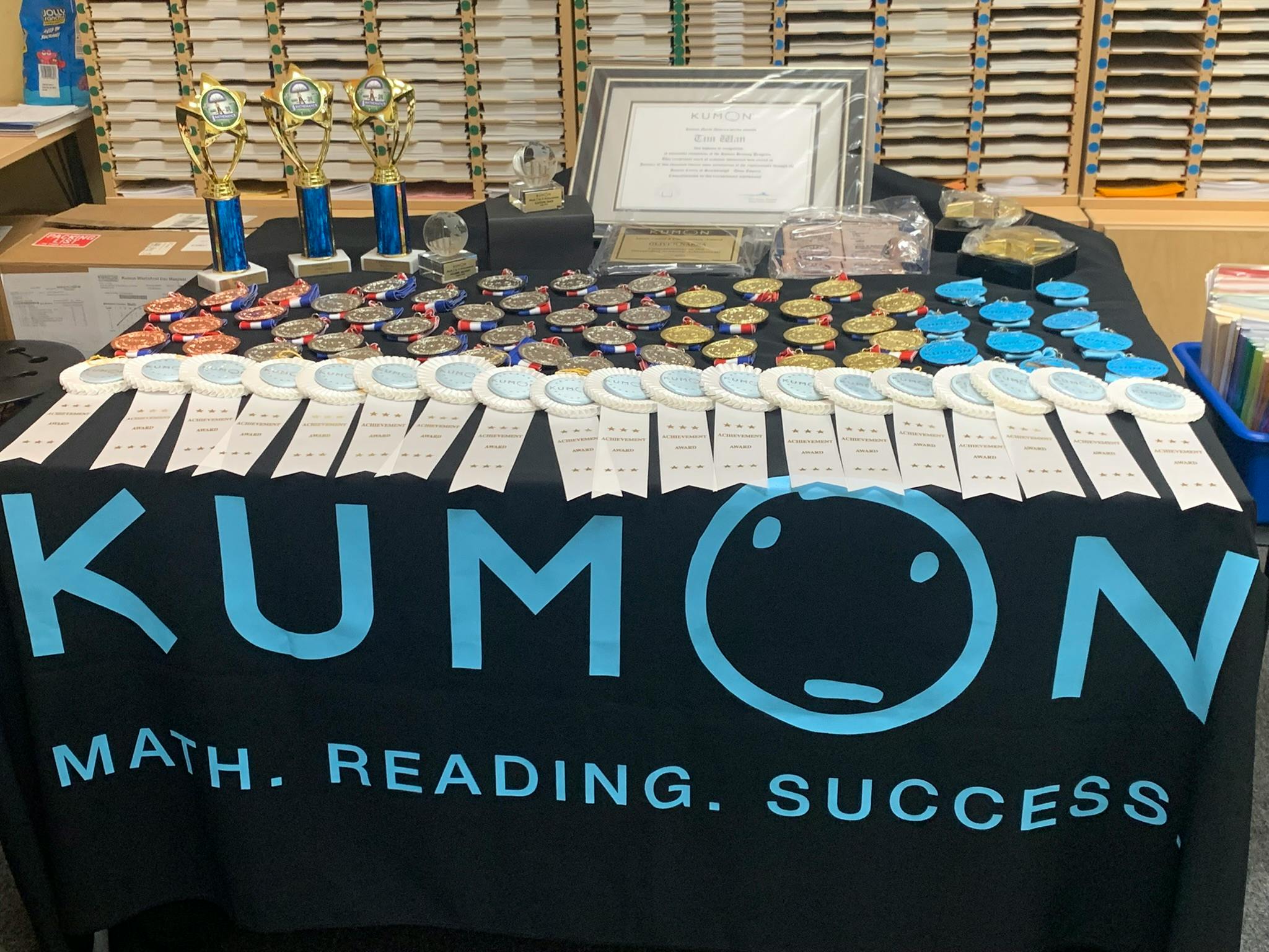 awards table with Kumon banner, trophies, medals, ribbons, and a certificate