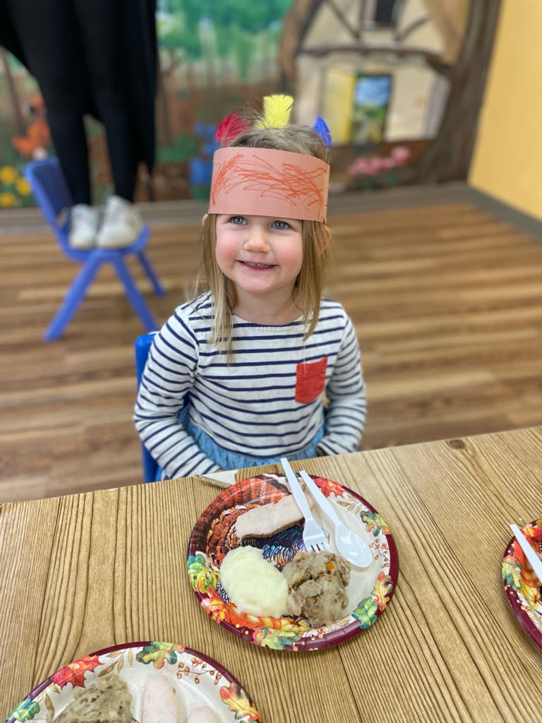 Child wearing a paper headband with feathers, sitting at a table with a plate of food