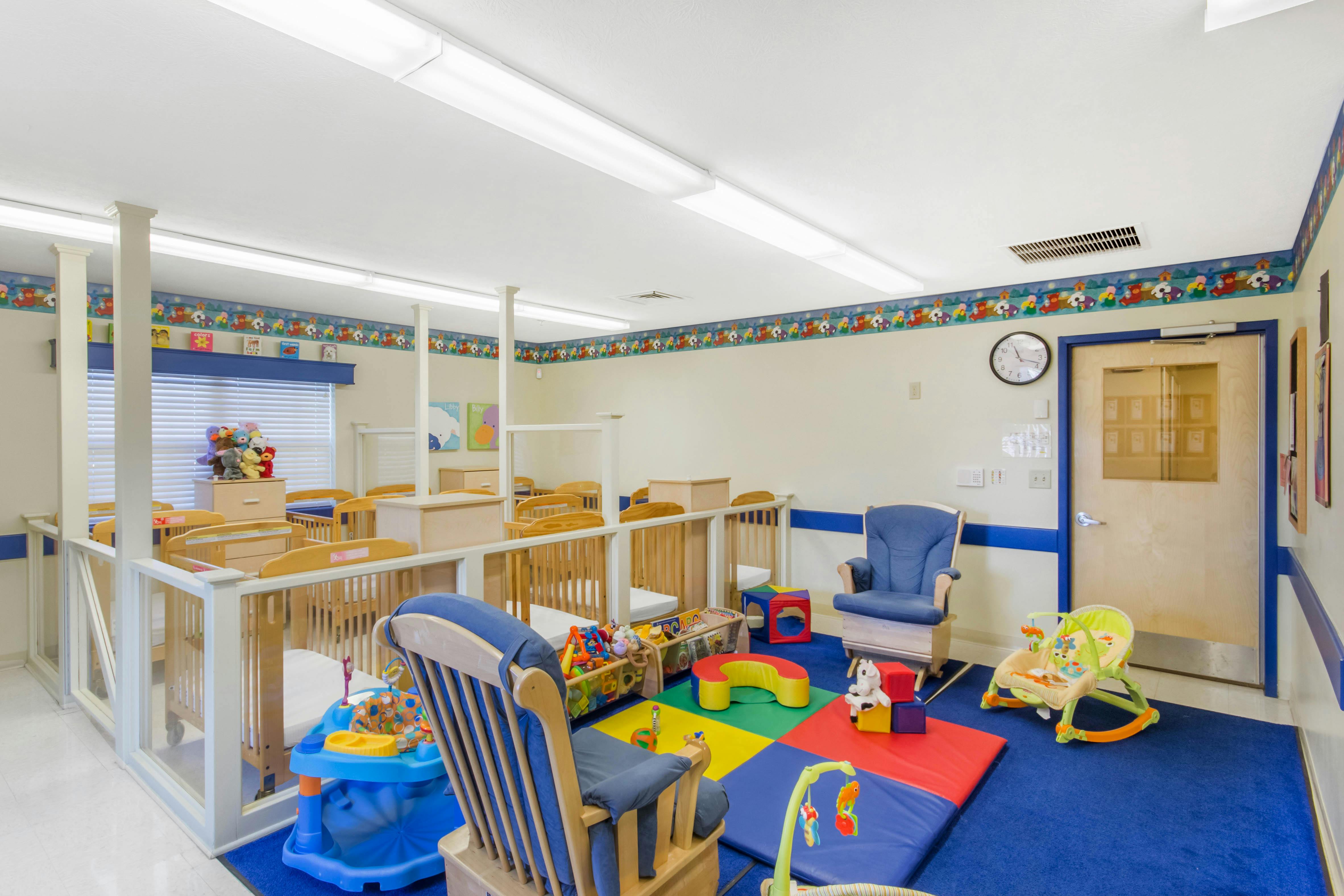 A colorful childcare room with cribs, play mats, and a blue rocking chair